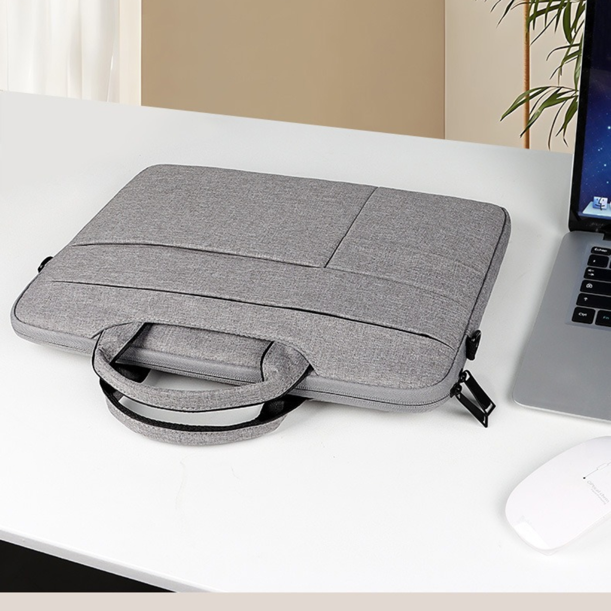 Graue Laptoptasche mit Griff auf einem weißen Schreibtisch, neben einem Laptop und einer weißen Maus. Im Hintergrund sind ein Fenster und eine Pflanze.