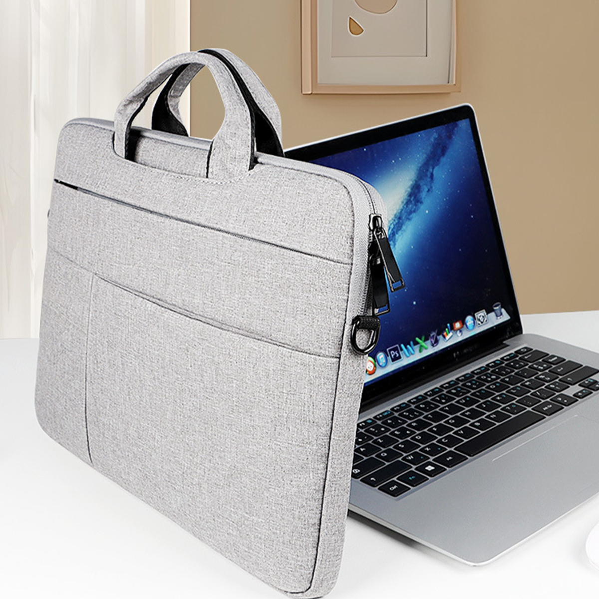 Graue Laptoptasche mit schwarzem Griff neben einem Laptop. Ein gerahmtes Bild ist im Hintergrund. Die Tasche steht auf einer weißen Oberfläche.