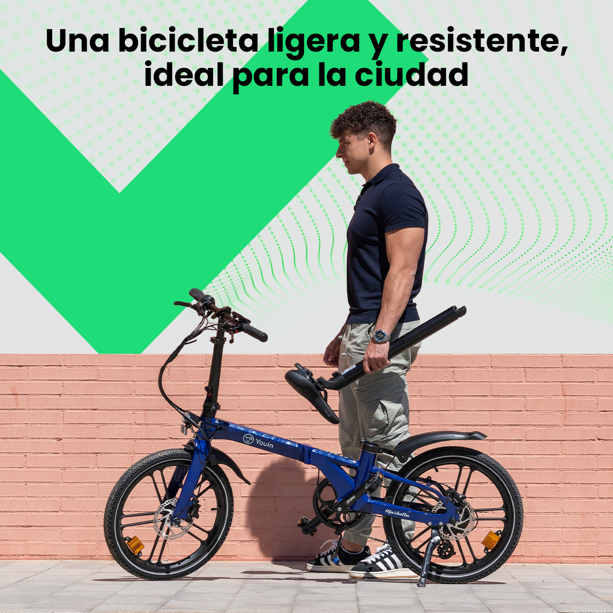 Un hombre sostiene una bicicleta negra plegada. Una bicicleta azul está delante. Una forma verde en una pared. Fondo de ladrillo rosa.