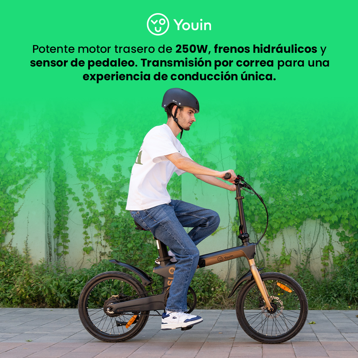 Hombre monta una e-bike Youin negra. Lleva casco, camiseta blanca y vaqueros. Pared verde al fondo.