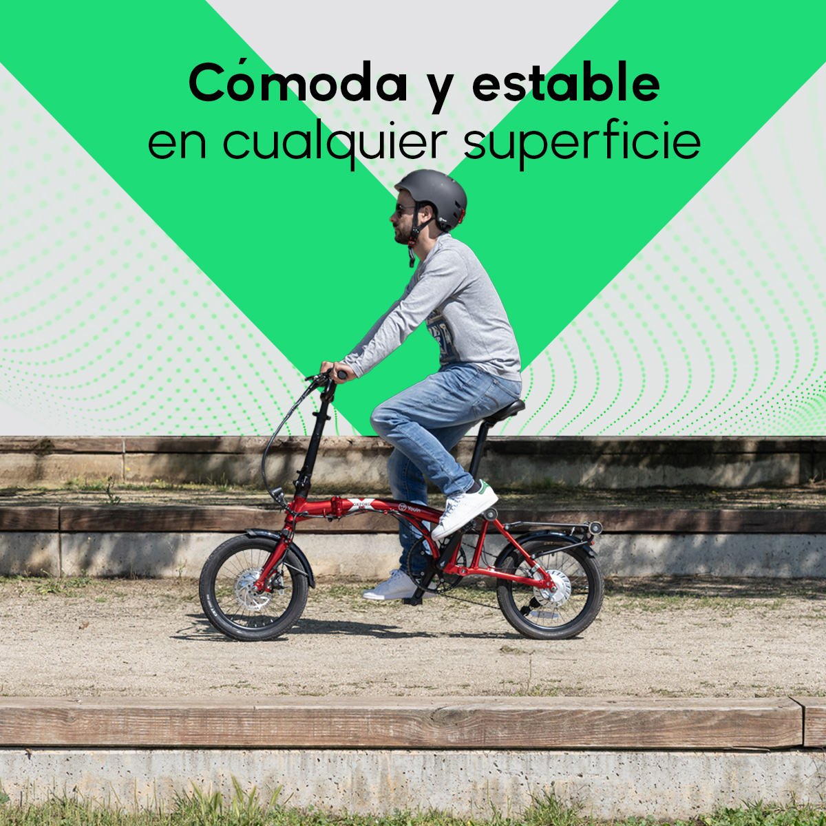 Hombre montando una bicicleta plegable roja en un camino. Lleva casco y vaqueros. El fondo son formas verdes y un parque.