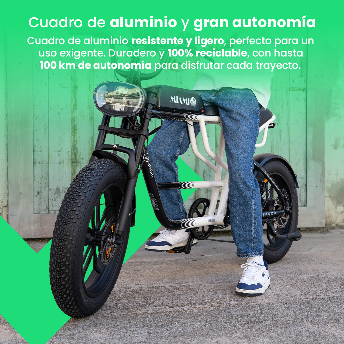 Persona en una bicicleta eléctrica. Llantas negras, marco blanco, jeans azules y zapatillas blancas. Puerta de madera verde en el fondo.
