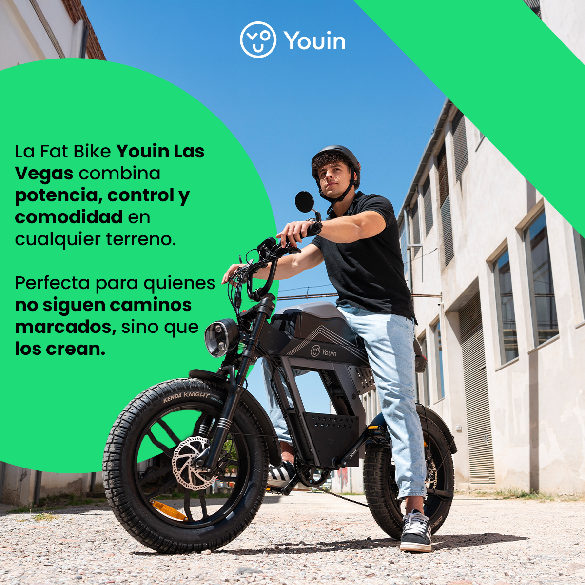 Hombre en bicicleta gorda negra. Lleva casco y vaqueros claros. El fondo es un edificio y cielo azul.