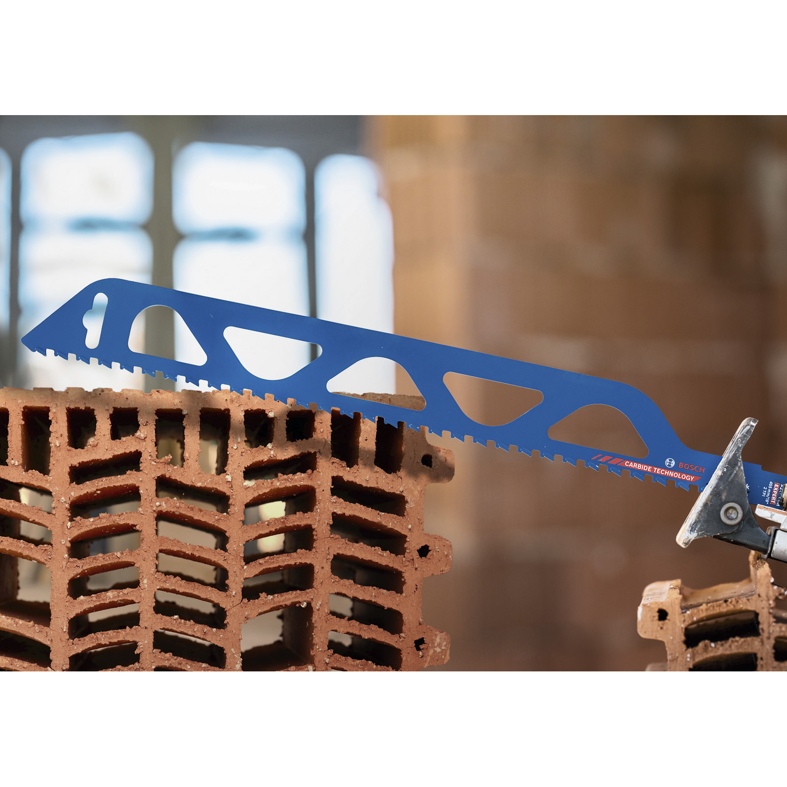 Niebieskie ostrze tnie brązową cegłę. Piła ma technologię węglikową. Tło ma rozmyte światło i cegły.