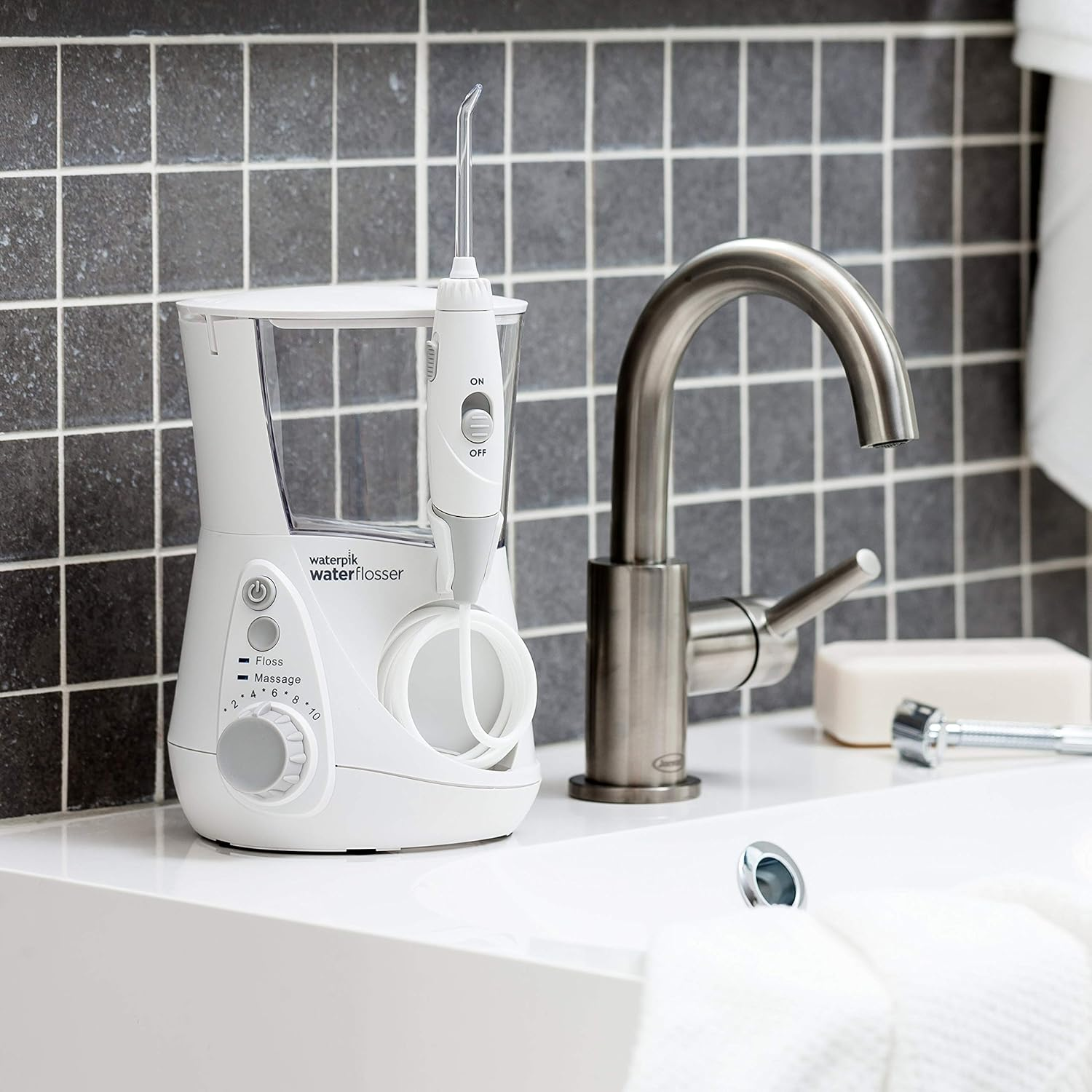 Un irrigador Waterpik blanco está sobre un lavabo blanco. Un grifo cromado está a su lado. El fondo está alicatado en gris.