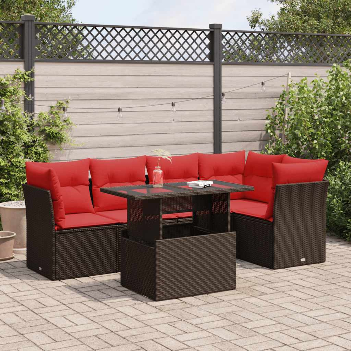 Terrassenmöbel-Set mit roten Kissen, braunen Korbmöbeln und einem Tisch mit Glasplatte. Hintergrund: Zaun und Grün.
