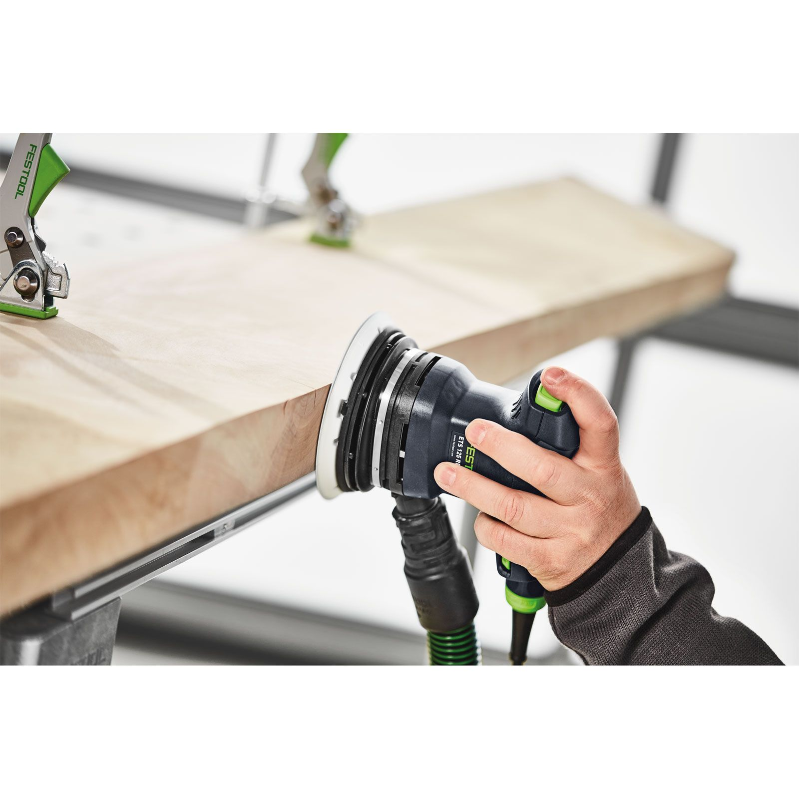 Eine Hand schleift eine Holzplanke mit einem Elektroschleifer. Der Schleifer ist schwarz und grün, das Holz hellbraun.