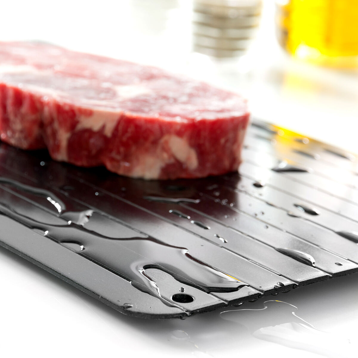 Eine schwarze Auftauschale mit Wassertropfen und einem gefrorenen Stück Fleisch darauf.