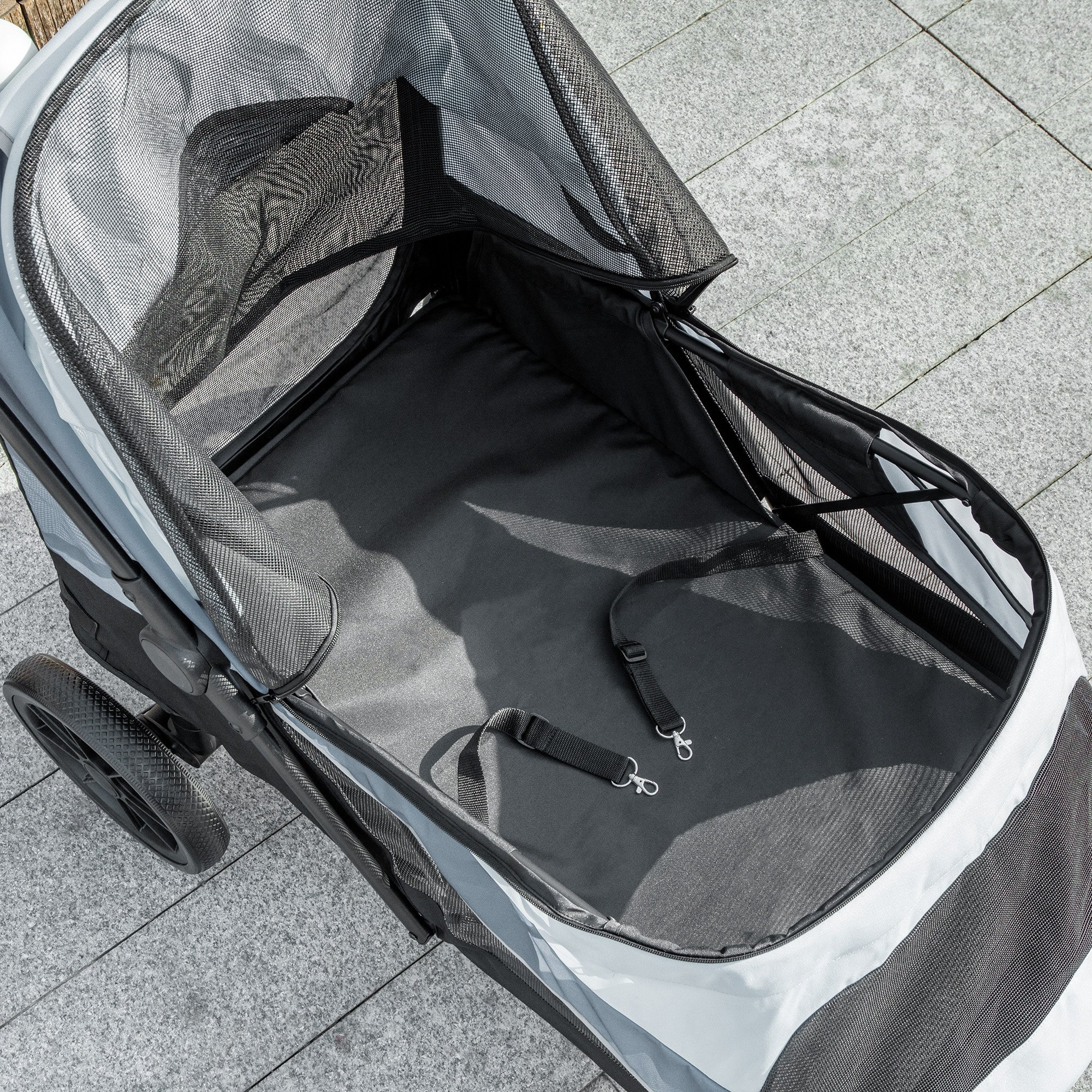 Ein offener Tierkinderwagen mit Mesh-Oberteil, schwarzem Innenraum und Sicherheitsgurten steht auf einer gefliesten Oberfläche.