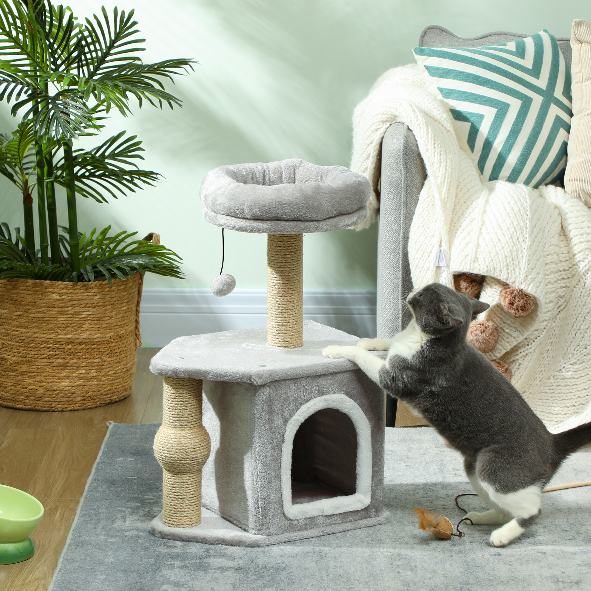 Un árbol para gatos gris con un gato de pie junto a él, con una planta y un sofá gris al fondo.