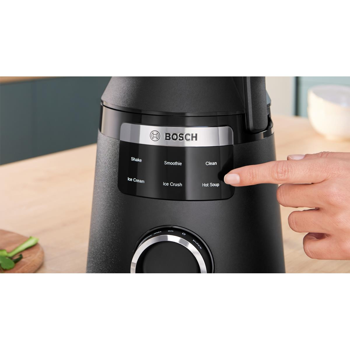 Frullatore Bosch nero con una mano che preme il pulsante "Zuppa calda", su una superficie di legno.