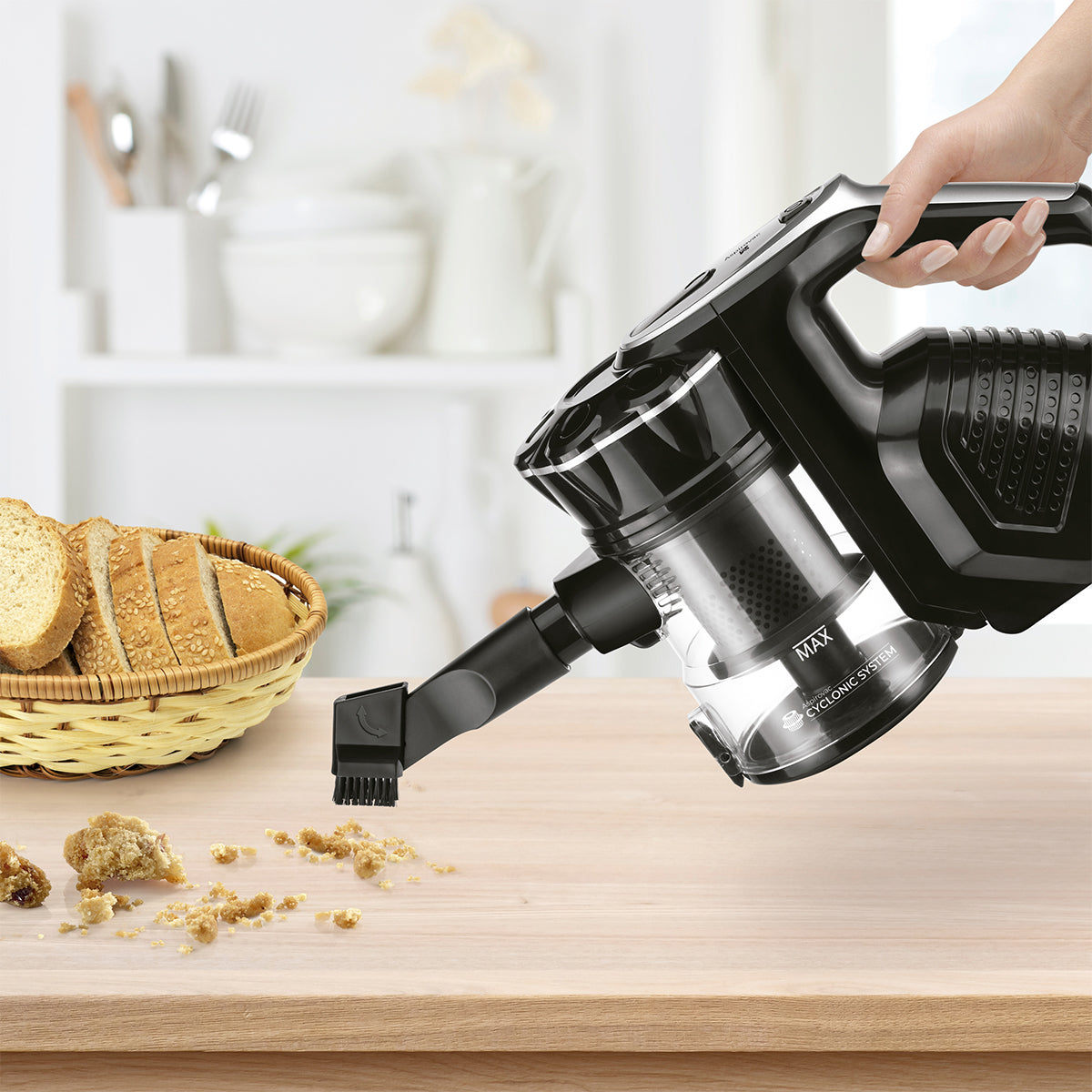 Una mano usa un aspirapolvere senza fili nero su un tavolo di legno, briciole e pane sono visibili.