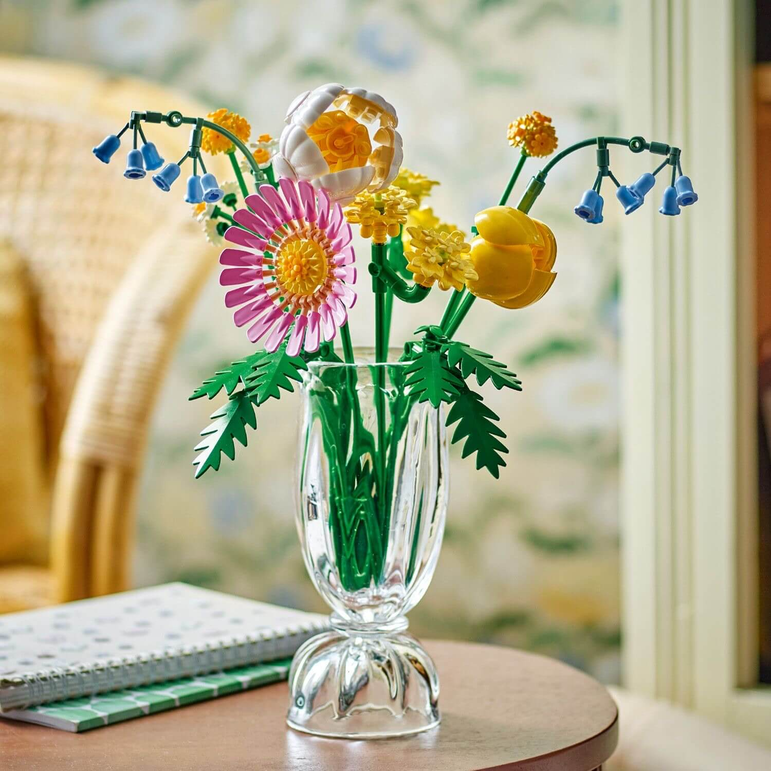 Bukiet kwiatów Lego w szklanym wazonie. Kwiaty są różowe, żółte, białe i niebieskie. Na stole leży notatnik.