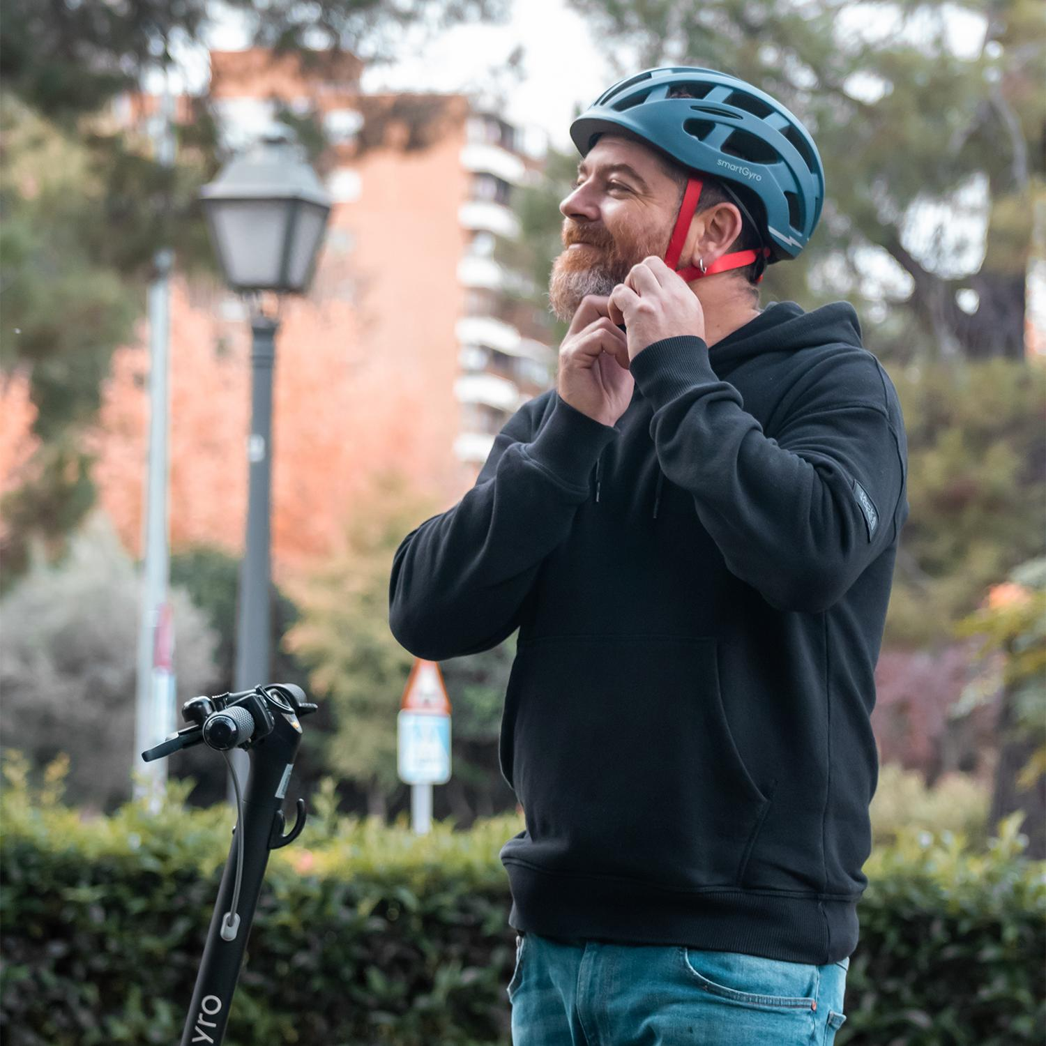Un hombre ajusta su casco junto a un scooter. Tiene barba y está al aire libre.