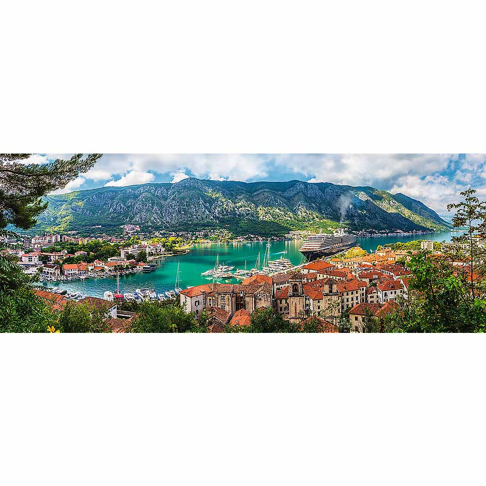 Vista panorámica de un pueblo costero, barcos, crucero, montañas, agua azul y edificios de tejados rojos, bajo un cielo nublado.