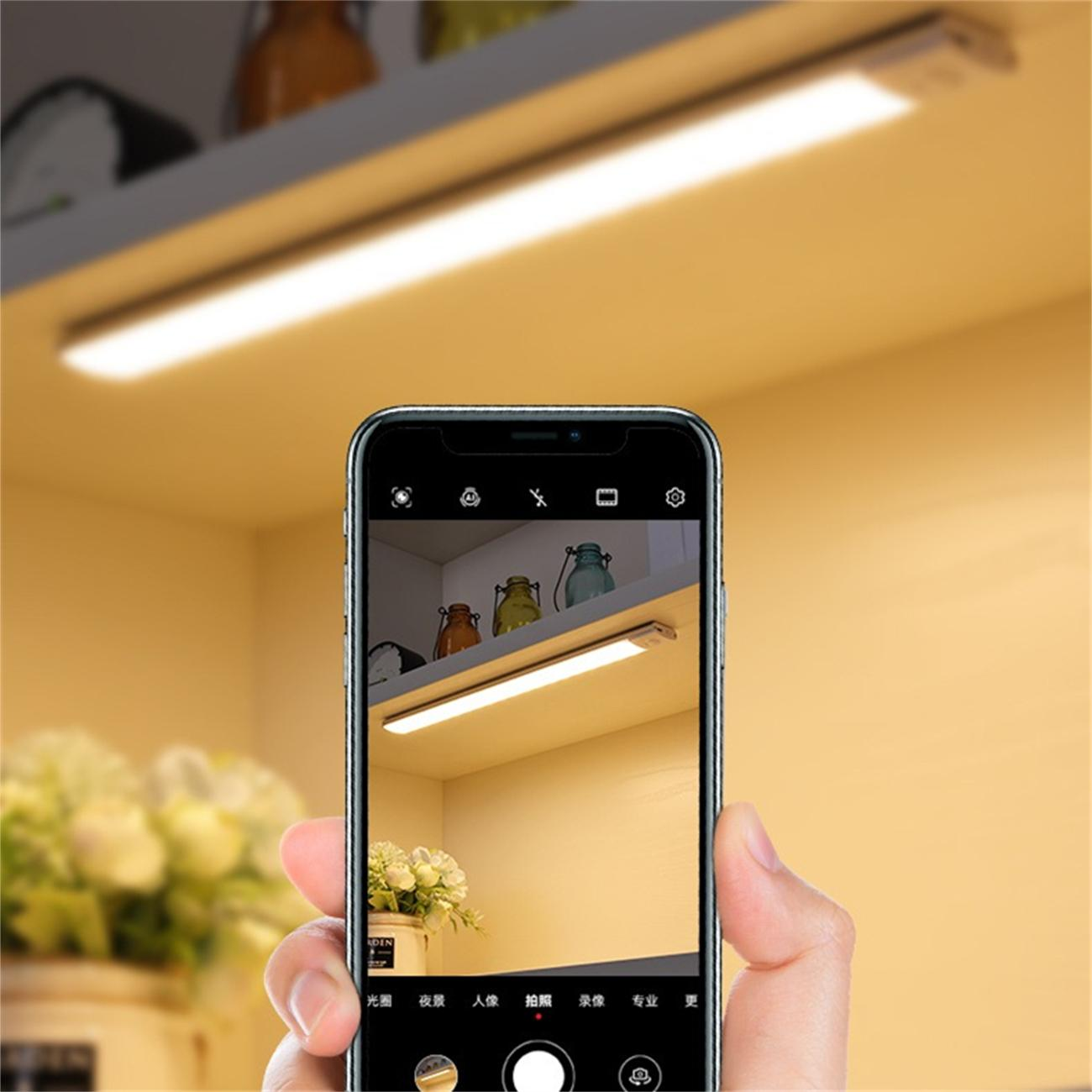 Eine Hand, die ein Telefon hält und ein Foto eines Lichtbalkens unter einem Regal mit Gläsern und Blumen aufnimmt.