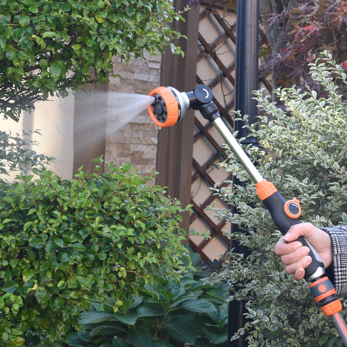 Eine Person benutzt einen Gartenschlauch mit orangefarbener Düse, um Wasser auf grüne Büsche in einem Garten zu sprühen.