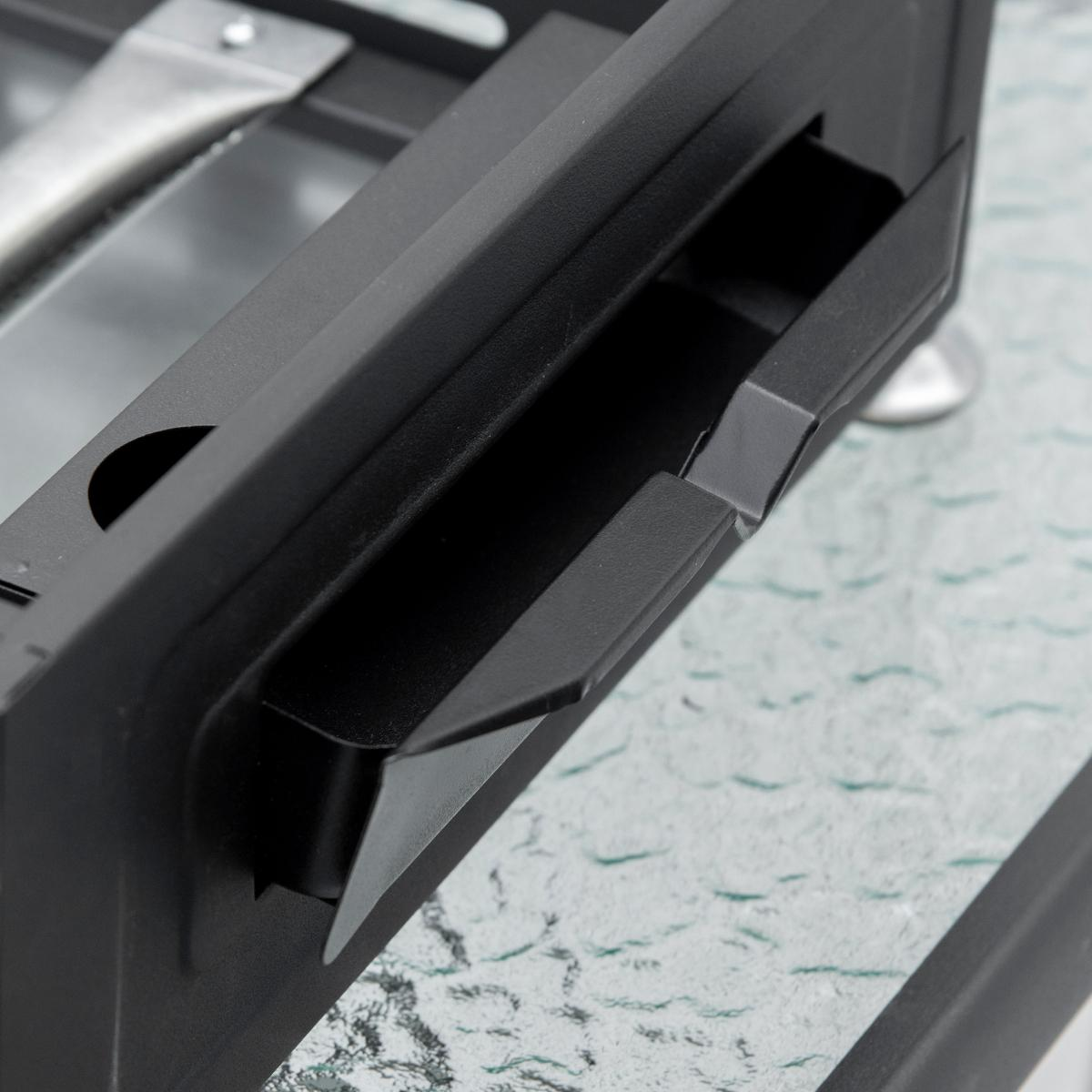 Nahaufnahme eines schwarzen Griffs an einem Grill. Der Griff ist Teil einer schwarzen Metallstruktur, die auf Glas ruht.