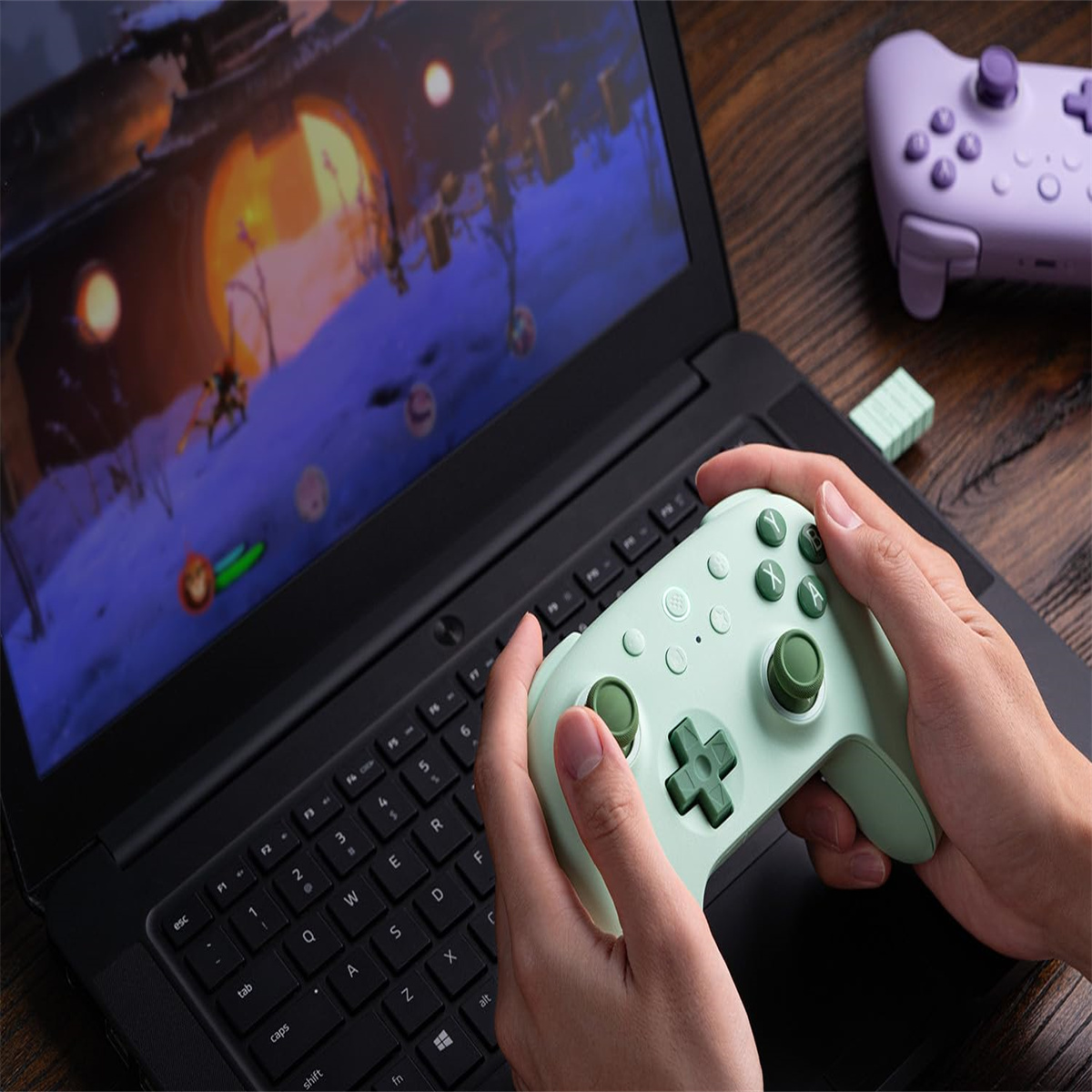 Eine Person hält einen grünen Controller vor einem Laptop, der ein Videospiel anzeigt. Ein weiterer Controller liegt auf dem Schreibtisch.