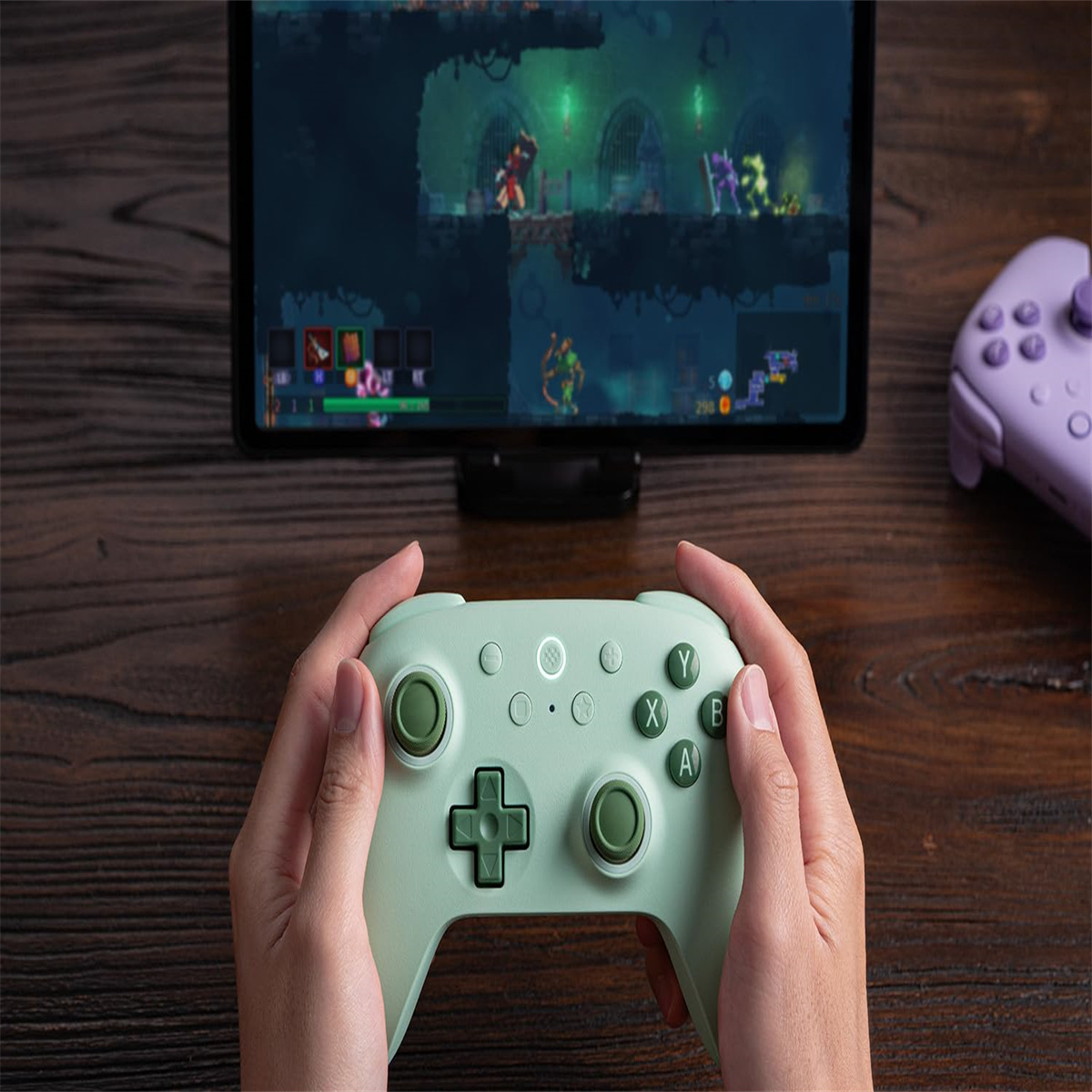 Hände halten einen grünen Gamecontroller und spielen ein Spiel auf einem Tablet. Ein lila Controller ist im Hintergrund.