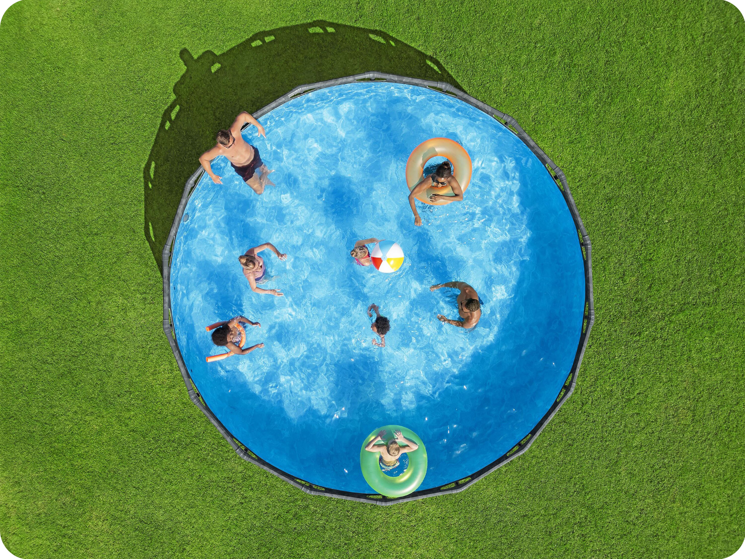 Widok z góry na osoby w okrągłym basenie. Zielona trawa otacza go. Niektóre osoby są w wodzie.