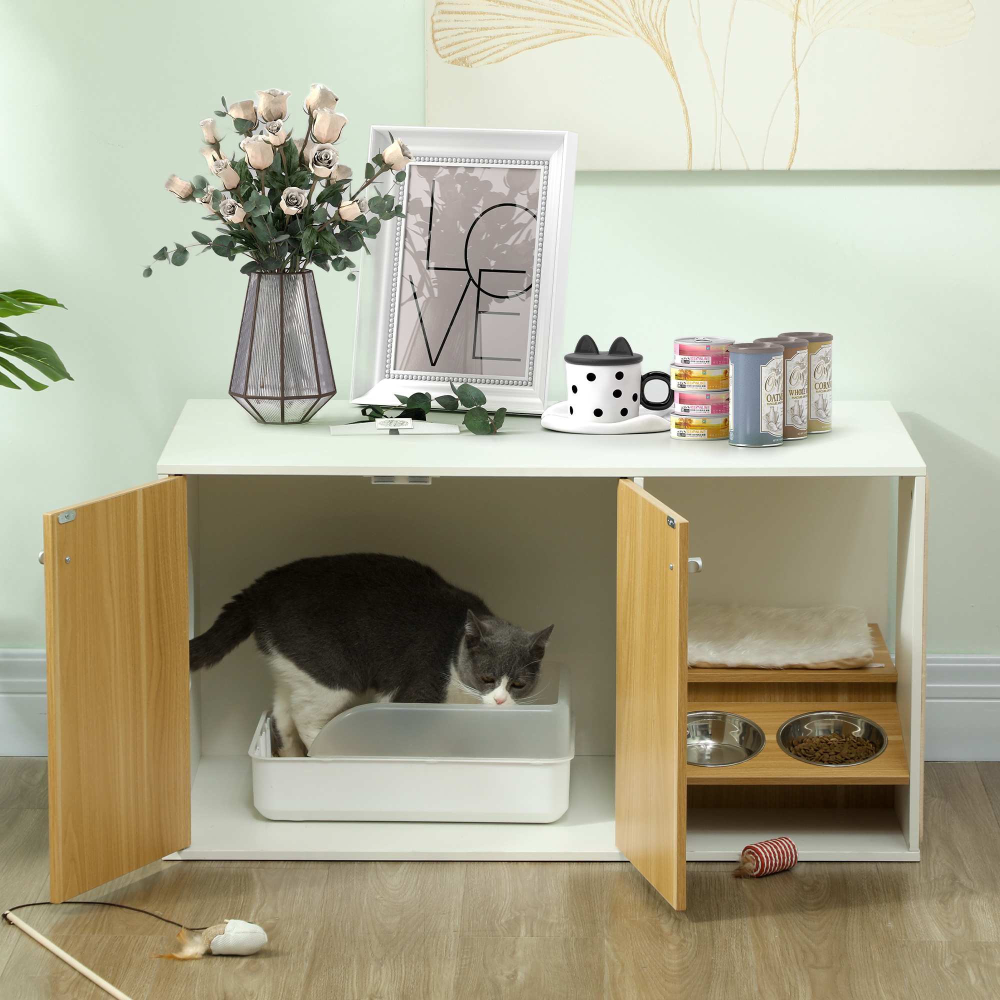 Una casa para gatos blanca y de madera con un gato usando la caja de arena y comederos.