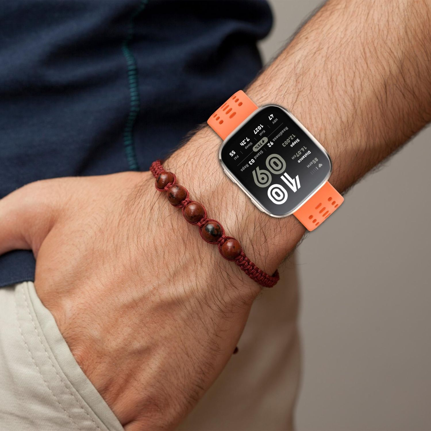 Handgelenk mit Smartwatch und braunem Perlenarmband. Die Uhr hat ein orangefarbenes Armband.