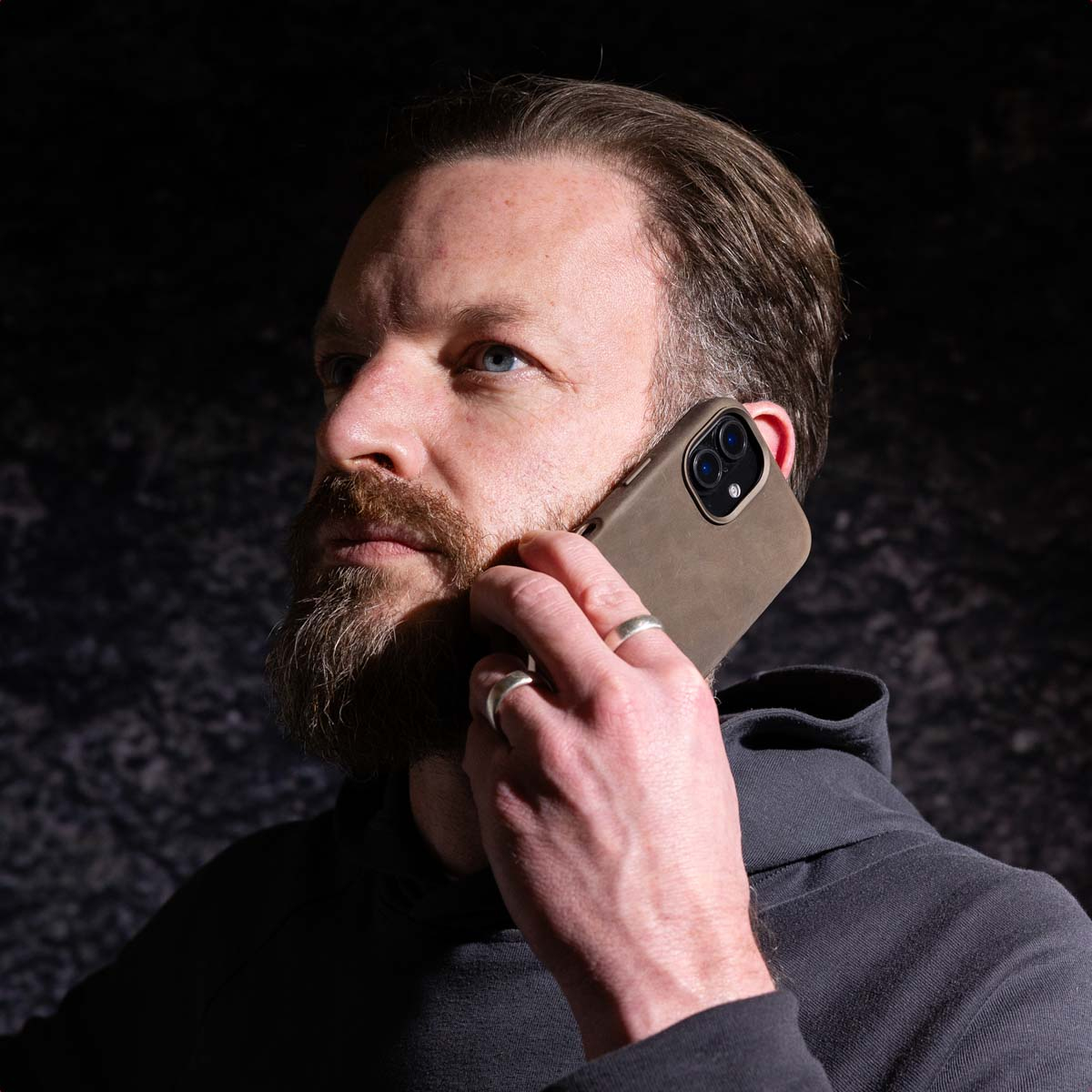 Un uomo con la barba porta un telefono all'orecchio. Indossa una felpa con cappuccio nera e un anello d'argento.