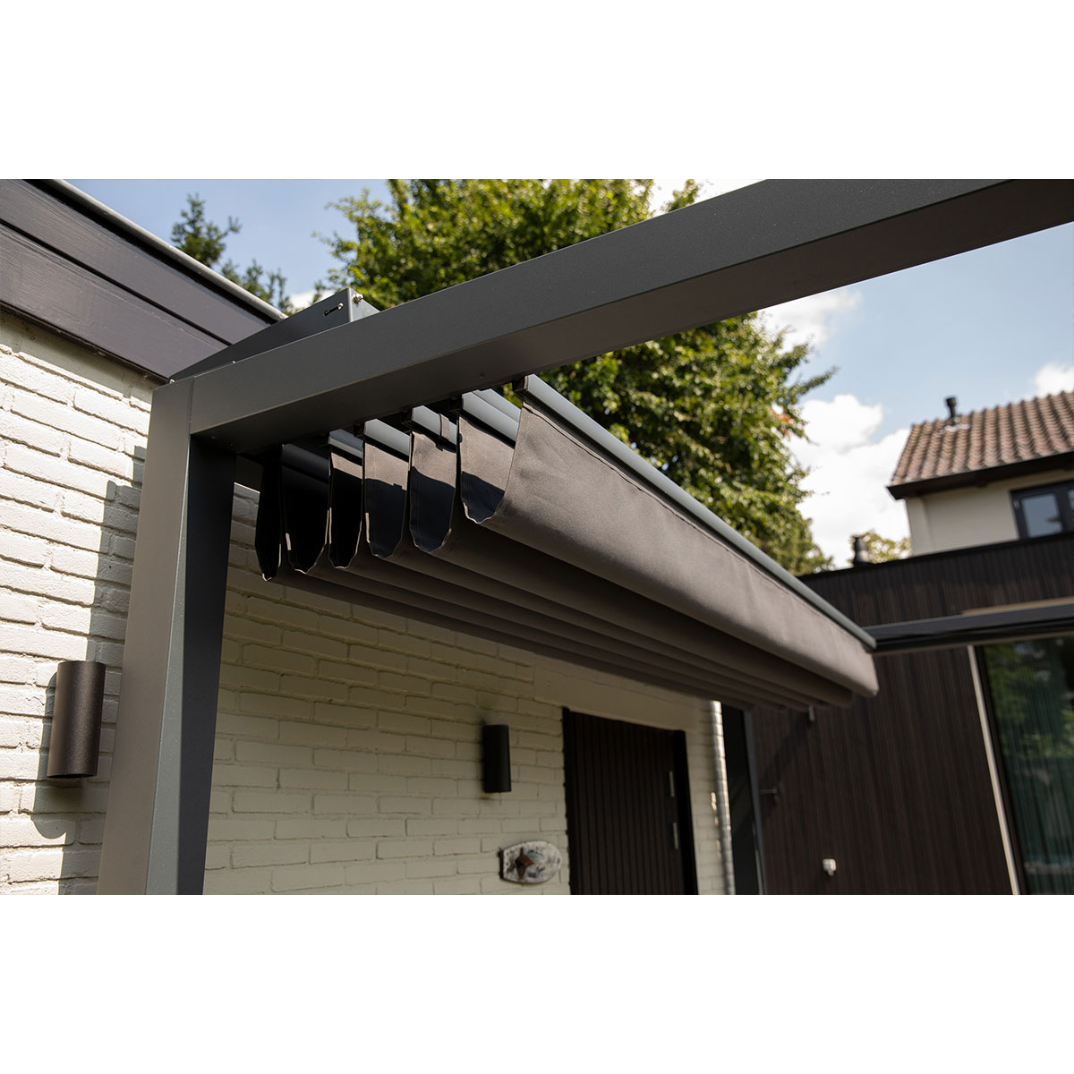 Pergola met grijs schaduwdoek en witte bakstenen muur, met een groene achtergrond.