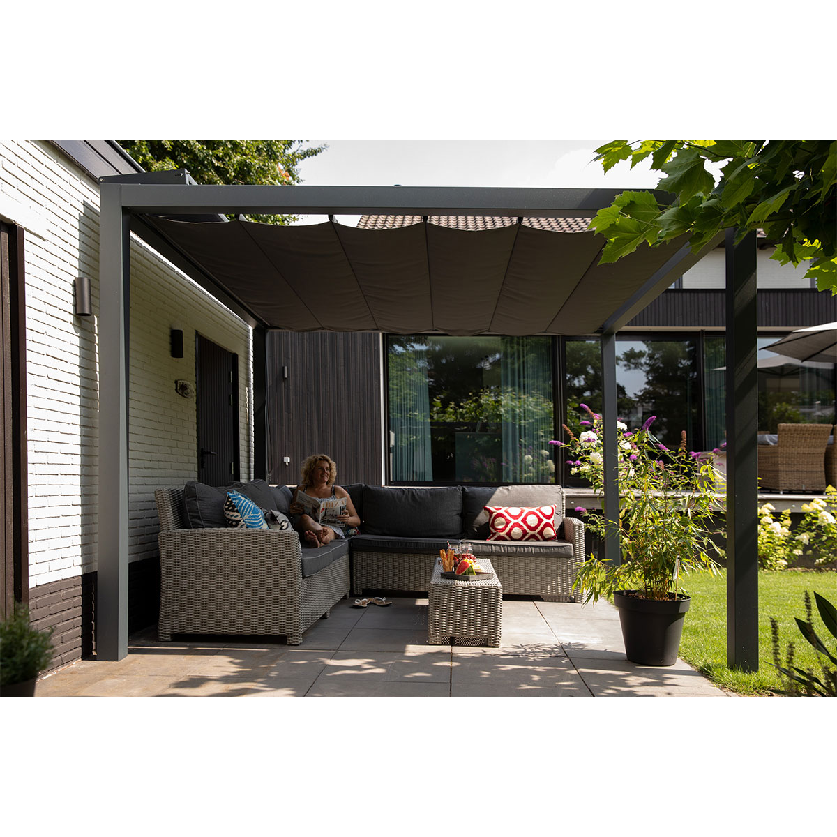 Een vrouw zit op een bank onder een pergola en geniet van drankjes. Planten en tuinmeubelen zijn zichtbaar.