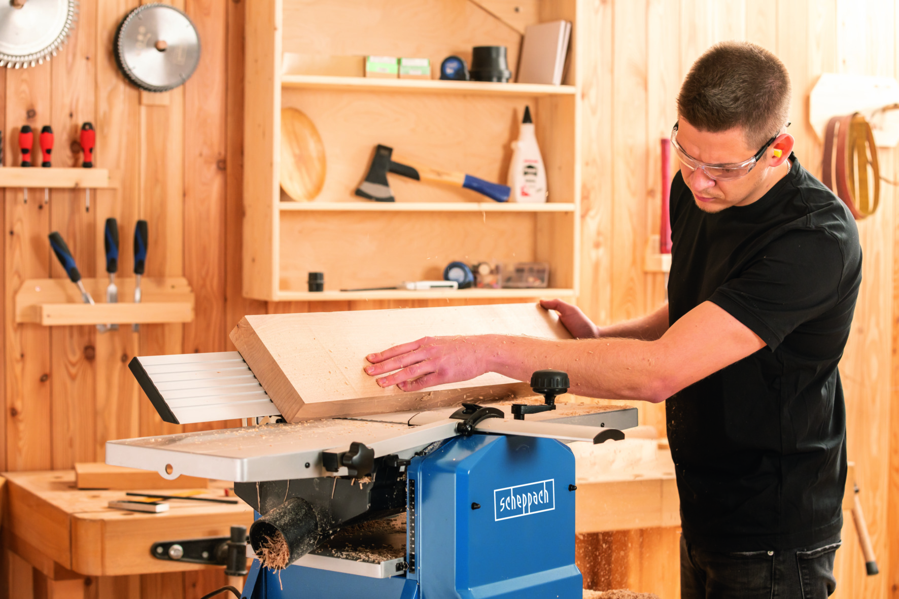 Ein Mann mit Schutzbrille bedient eine blaue Hobelmaschine. Er hobelt ein Holzbrett in einer Holzwerkstatt.
