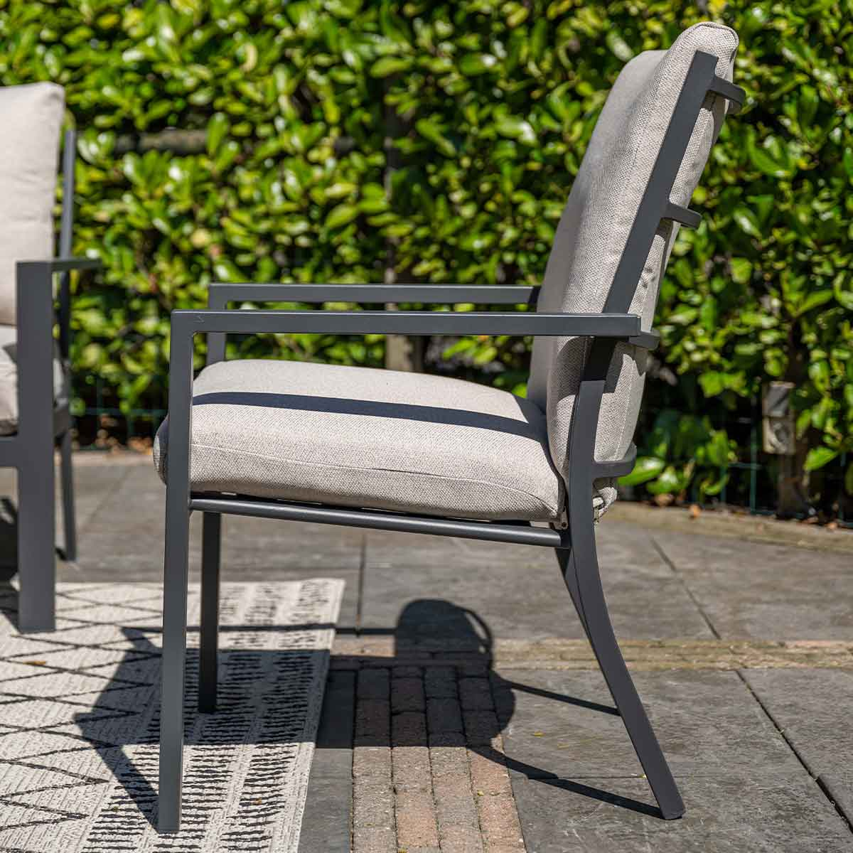 Grijze buitenstoel met wit kussen. De stoel staat op een stenen terras met groene struiken op de achtergrond.