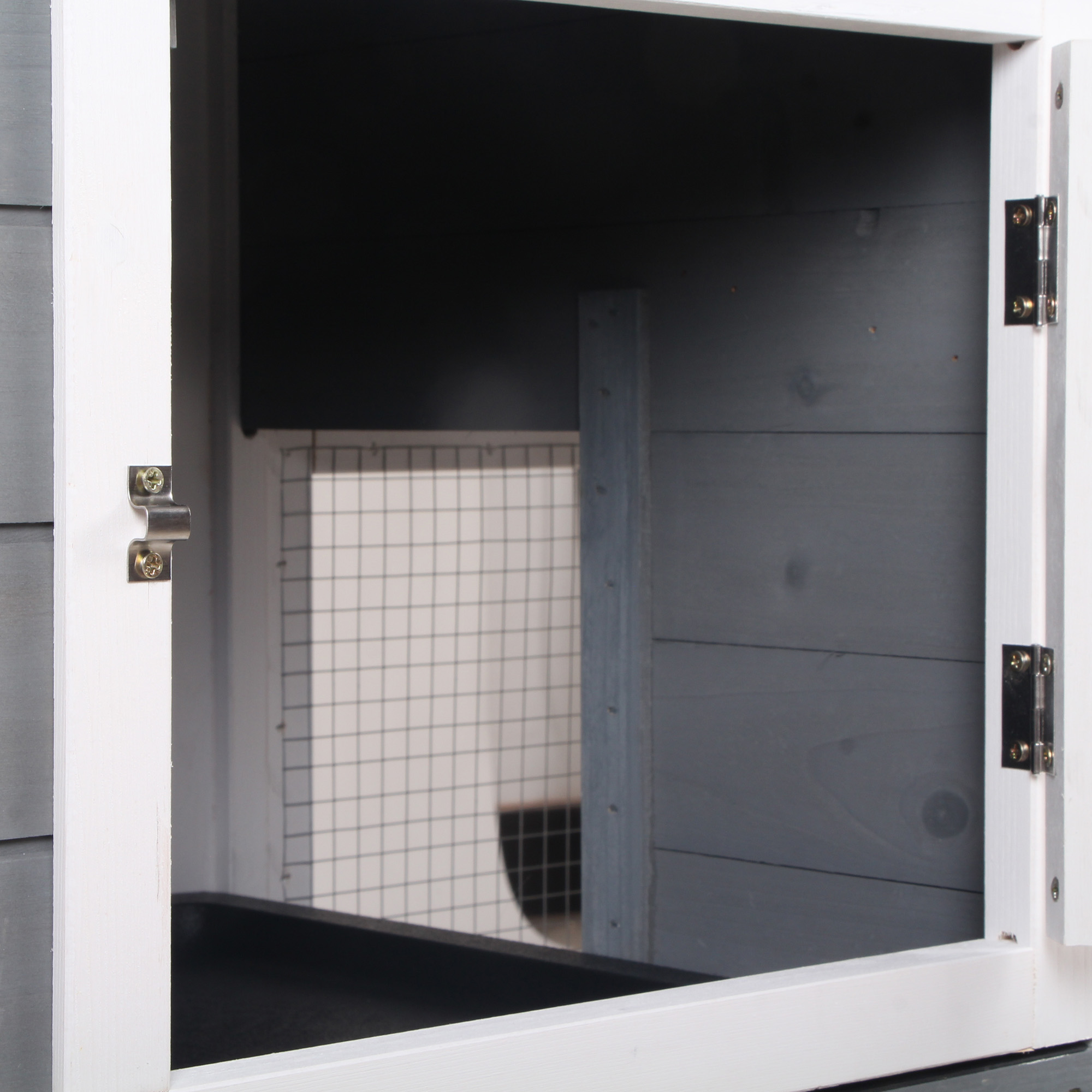 La puerta abierta de una casa para mascotas muestra un interior de malla metálica, piso oscuro y paredes grises, vista desde afuera.
