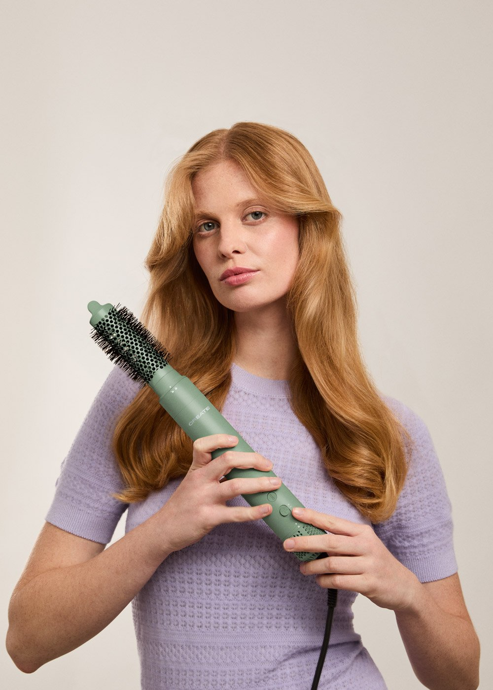 Una mujer pelirroja sostiene un cepillo verde para peinar el cabello. Lleva un suéter morado, mirando a la cámara.