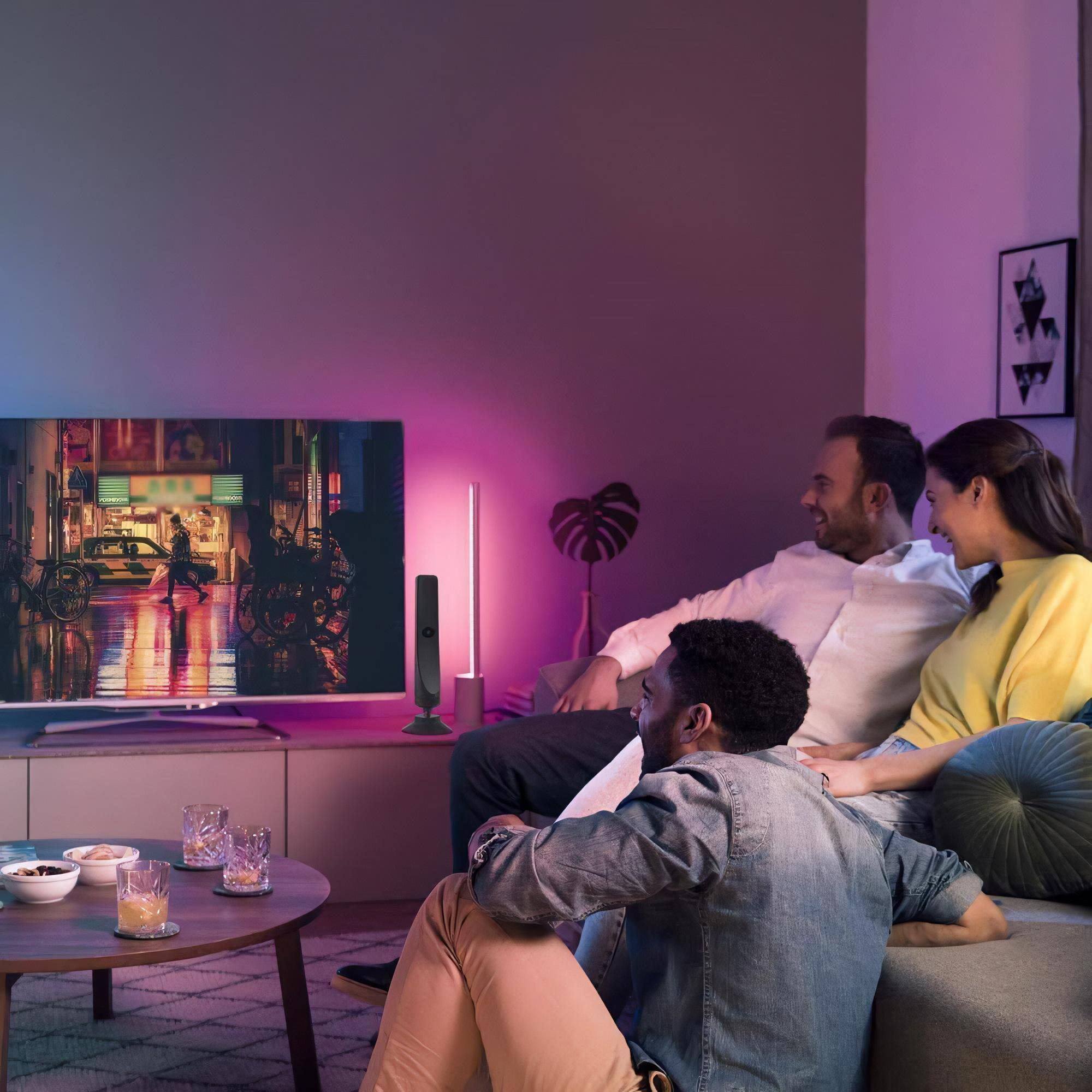 Drei Personen auf Couch beim Fernsehen, Tisch mit Getränken. Lila und rosa Ambientebeleuchtung schafft gemütliche Atmosphäre.