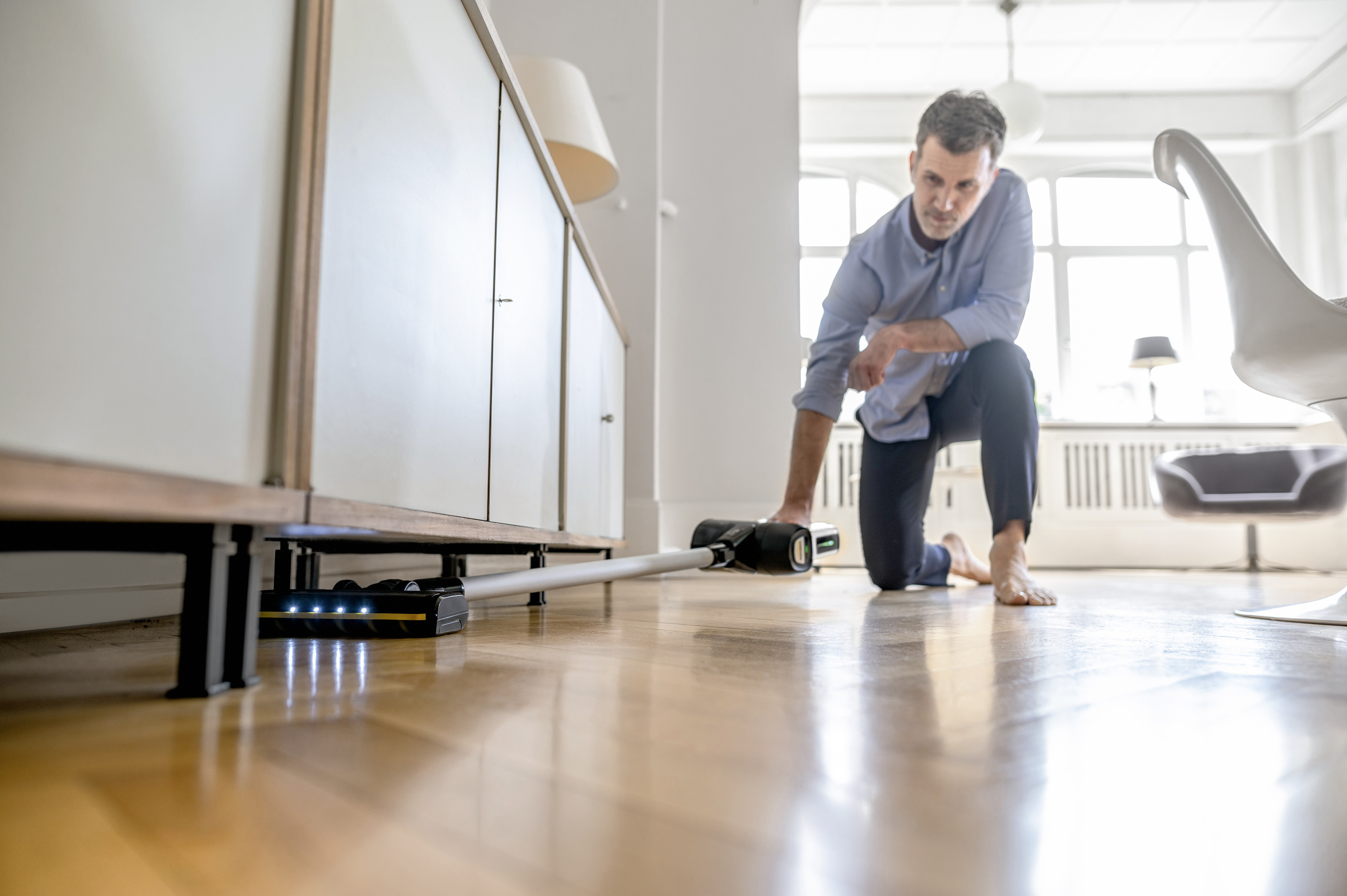 Un uomo sul pavimento che passa l'aspirapolvere. Un pavimento in legno è visibile e un mobile bianco è a sinistra. Una lampada è a sinistra.