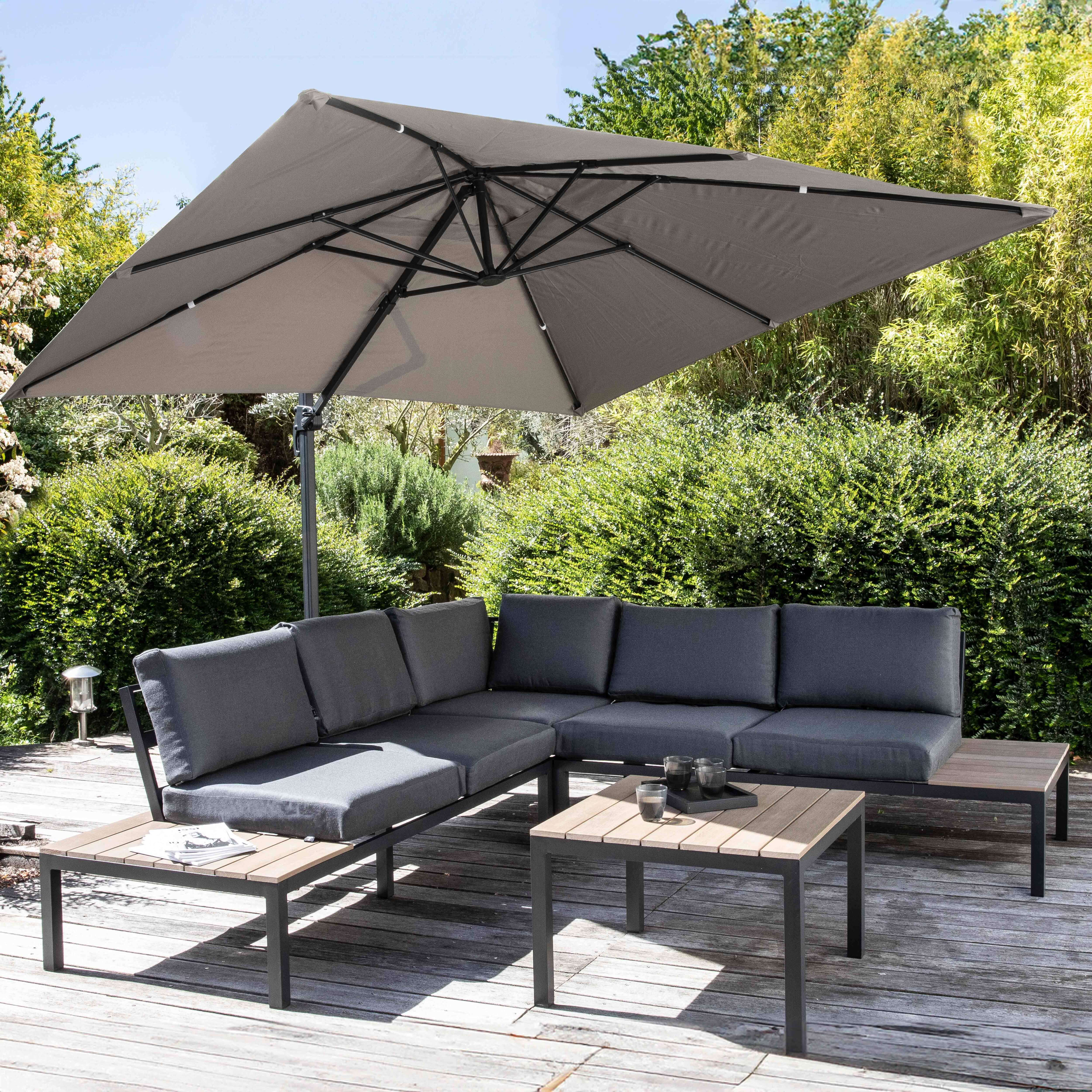 Outdoor-Patio-Setting mit grauem Ecksofa, zwei Tischen und einem offenen quadratischen Schirm. Grüne Büsche im Hintergrund.