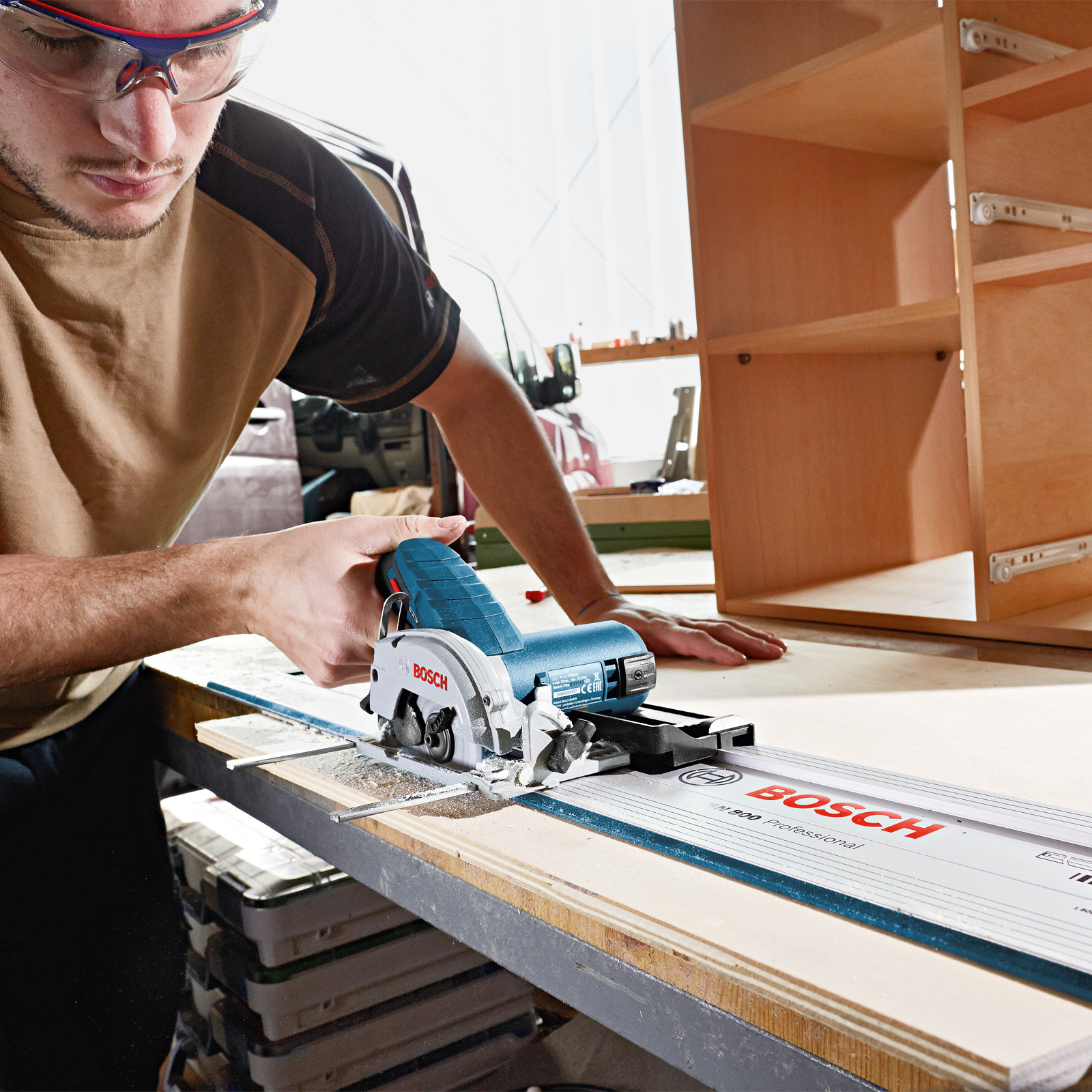 Mężczyzna używa niebieskiej piły tarczowej Bosch do cięcia drewnianej deski, w okularach ochronnych, z szafką w tle.