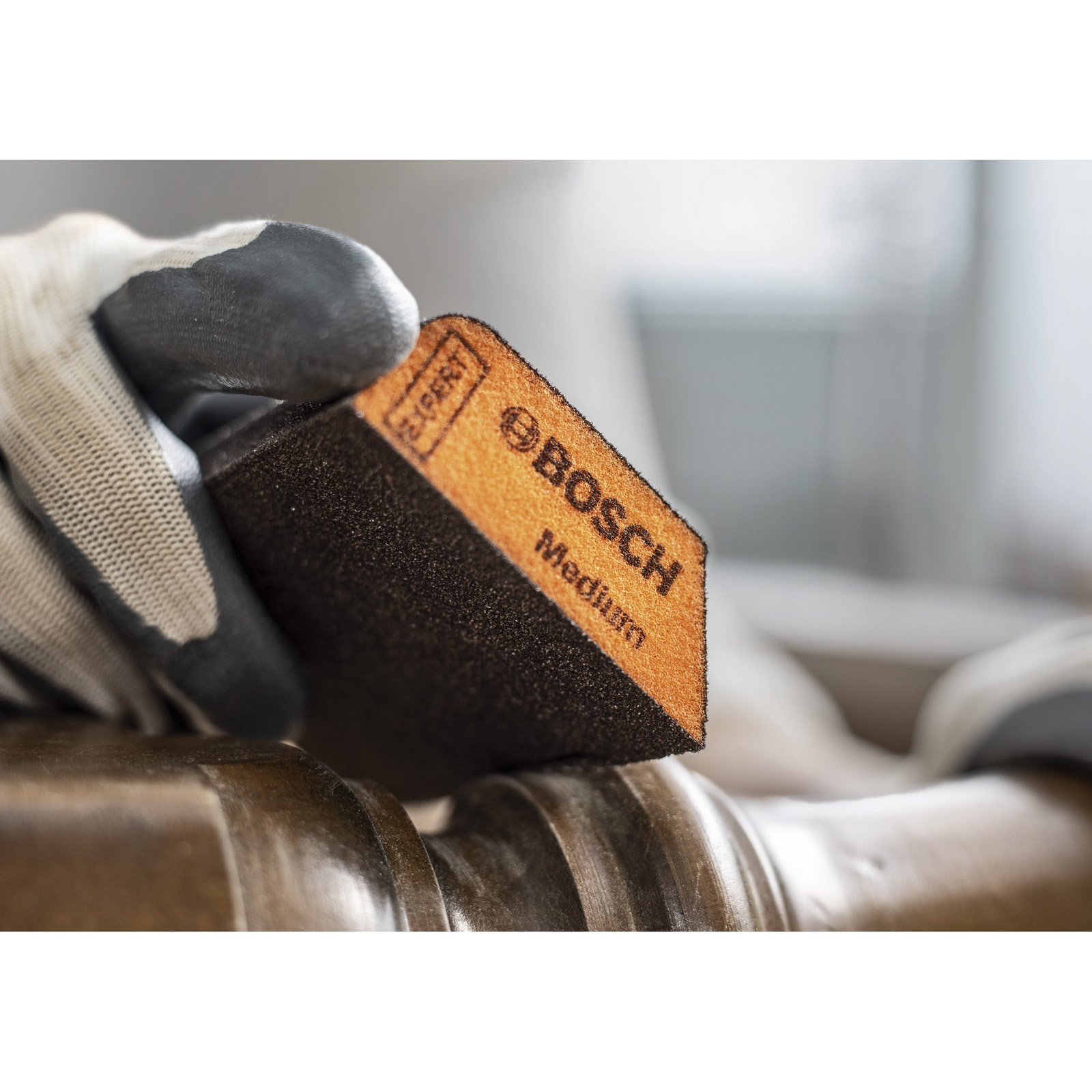 Eine behandschuhte Hand hält einen schwarzen Schleifklotz mit dem Bosch-Logo. Der Klotz ist braun mit schwarzem Sockel.