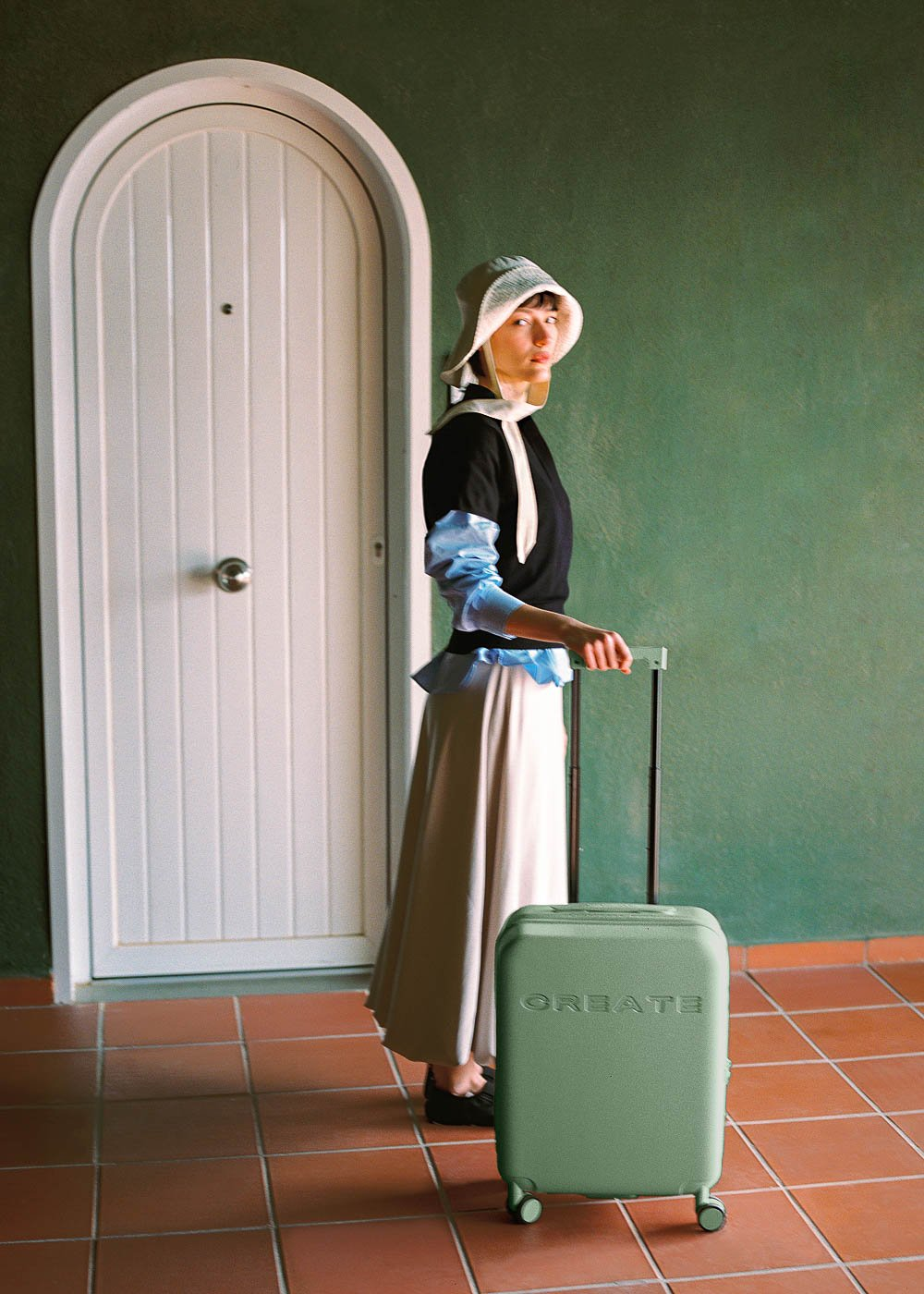 Mujer con sombrero y maleta verde, de pie cerca de una puerta blanca y pared verde.