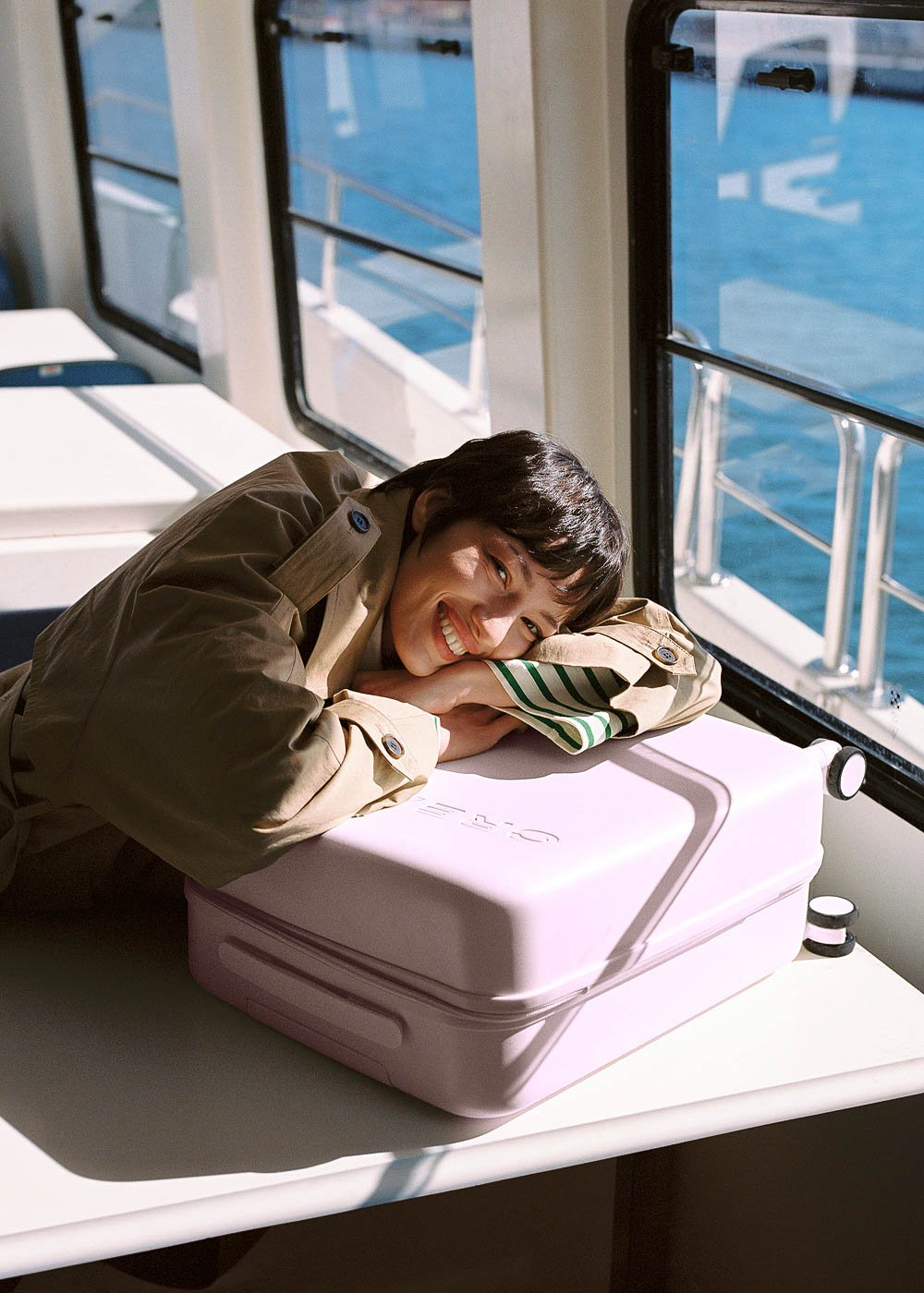 Una persona con un abrigo beige sonríe, apoyada en una maleta lavanda 'CREATE' en la cabina de un barco, con una ventana brillante detrás.