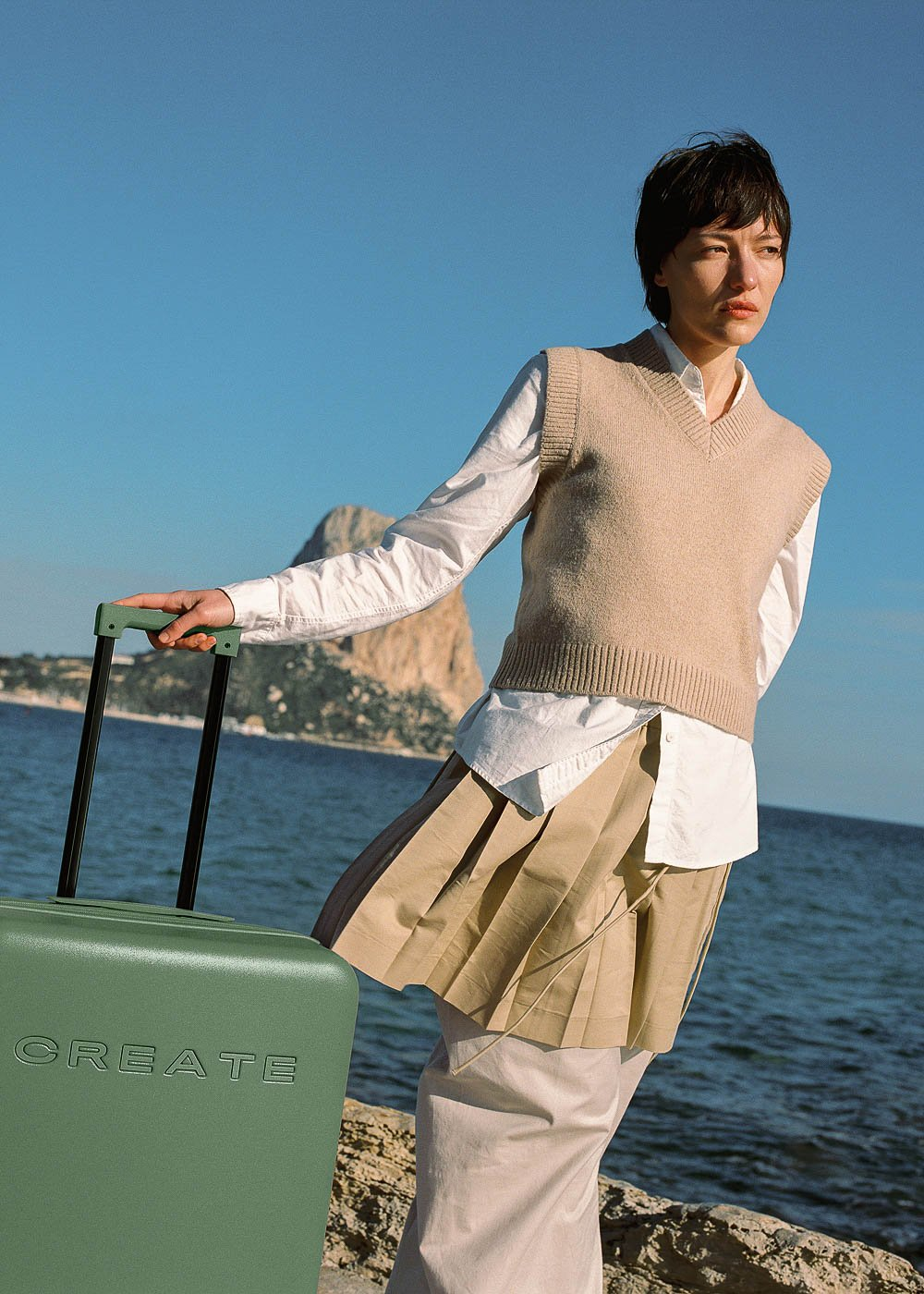 Una mujer con una maleta verde junto al mar, con chaleco beige y camisa blanca.