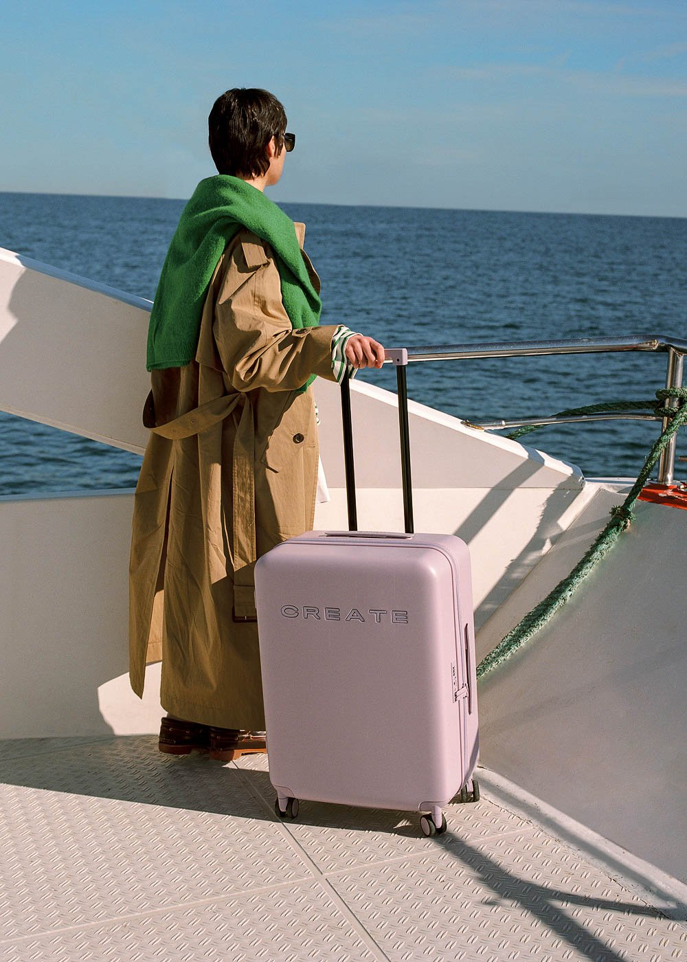 Una persona con abrigo y suéter verde en un barco con maleta CREATE lila, mirando el mar.