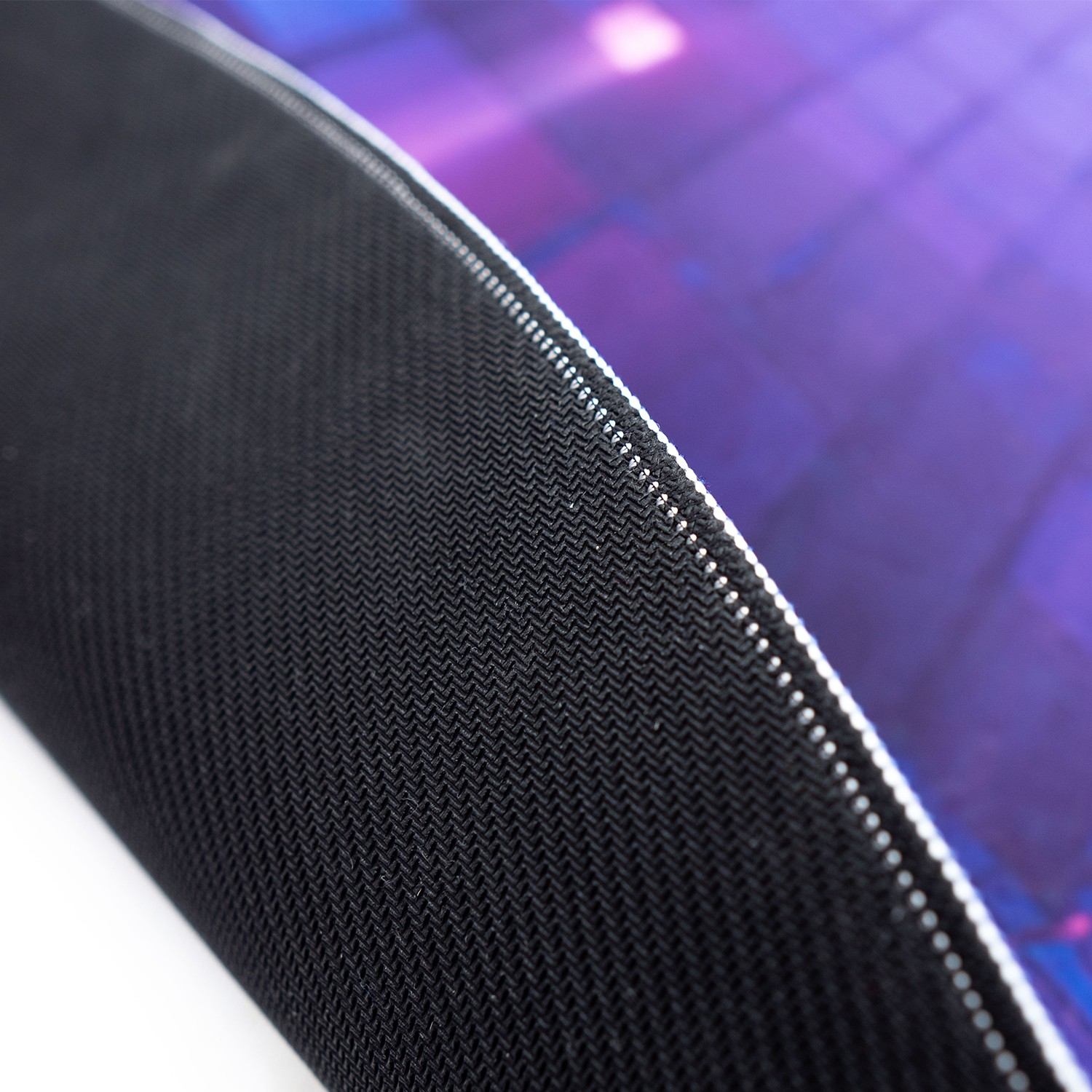 Primer plano de una alfombrilla gaming negra. Parte inferior visible, con tejido texturizado. Gráfico morado/azul.