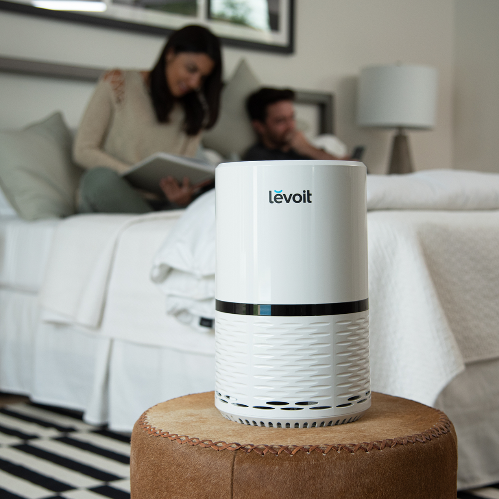 Oczyszczacz powietrza na stołku w sypialni; para czyta książkę i korzysta z telefonu.