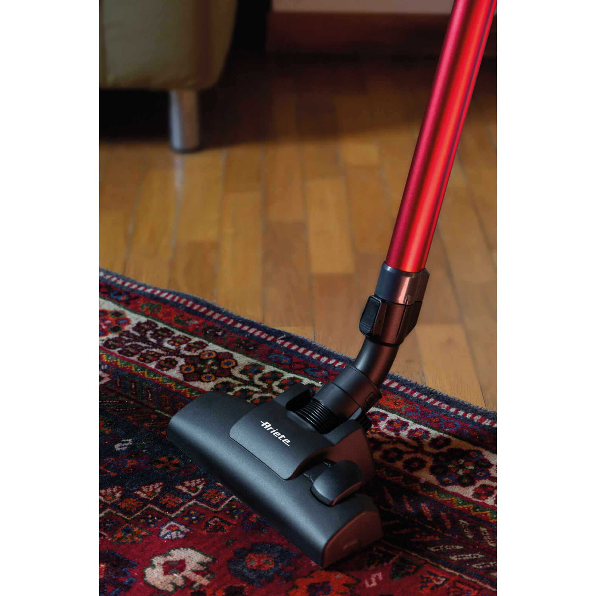 Czerwony odkurzacz na wzorzystym dywanie. Dywan leży na drewnianej podłodze. W tle widoczna jest sofa.