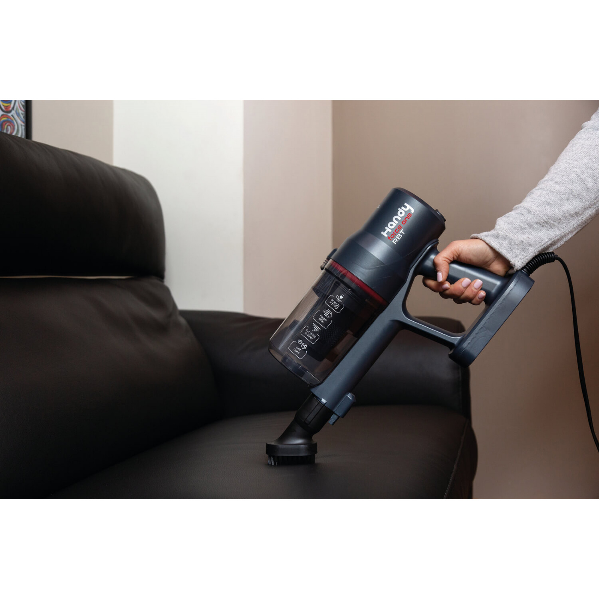 Osoba używająca odkurzacza ręcznego na czarnej sofie. Odkurzacz jest granatowy z czerwonymi akcentami.