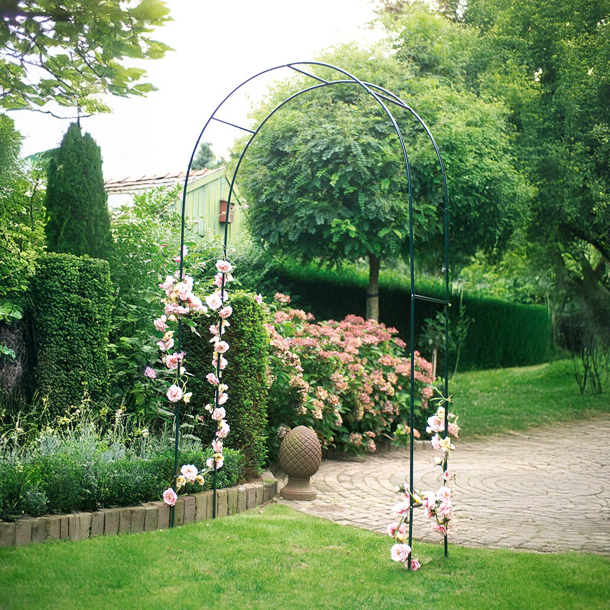 A garden arch with pink flowers and greenery. The arch is metal. Bushes and trees are in the background.