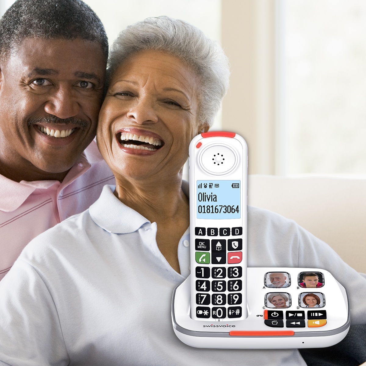 Un couple de personnes âgées sourit, un téléphone devant eux. Le téléphone est blanc avec de gros boutons. Il y a une ligne rouge en haut.