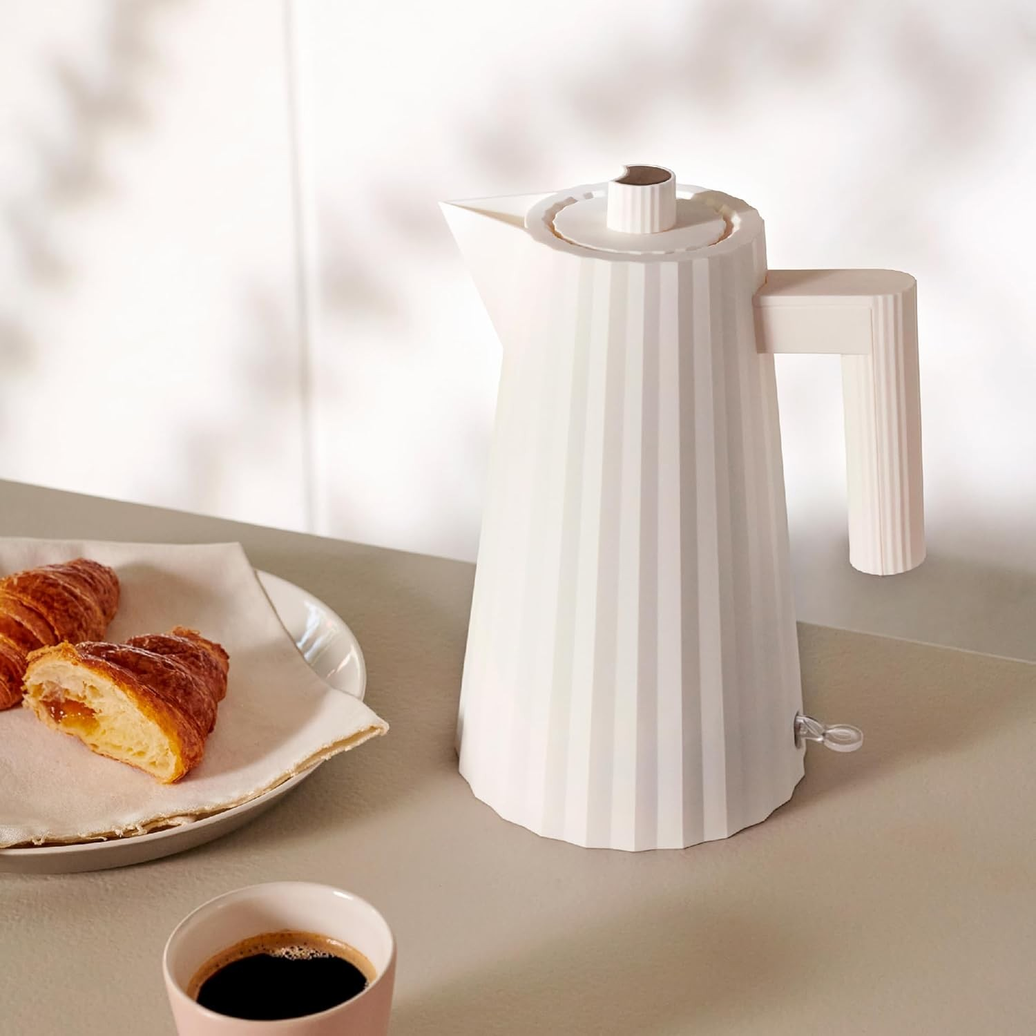 Bollitore elettrico bianco, croissant su un piatto e una tazza di caffè su un tavolo. Sfondo luminoso.